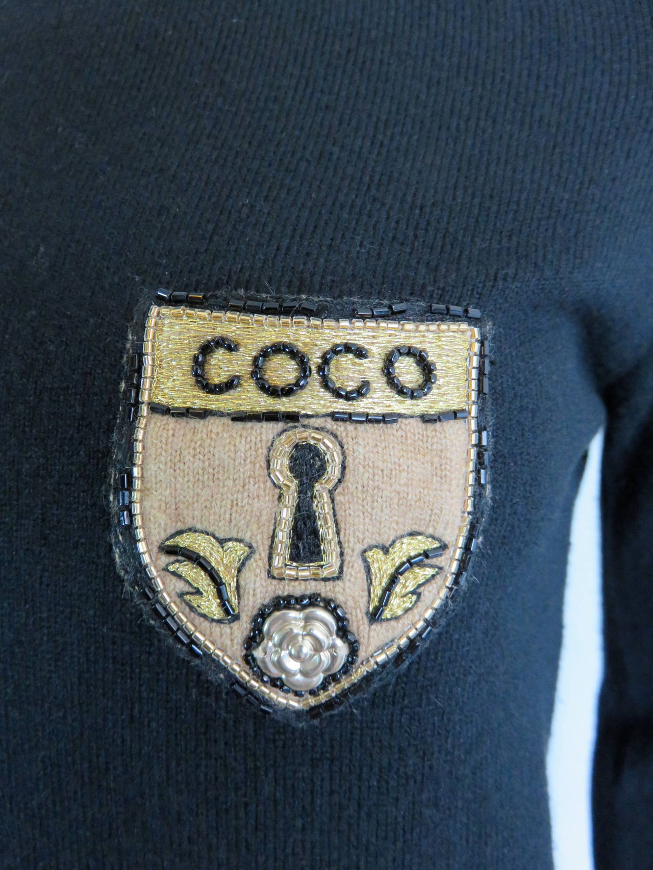 Excellent condition, CHANEL PARIS, pure cashmere sweater with embellished 'COCO' shield patch at the left chest.  

Crew-neck style sweater in black with beige contrast rib at neckline, and dual cuffs.

The matching, cashmere knit patch is