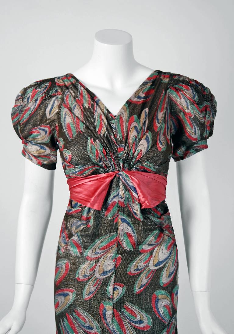 A rare luxurious atomic-deco print shimmer lame gown from the "Old Hollywood" era of glamour. The bodice is a dramatic pintuck V-neck with stylized short puff-sleeves. The waist is nipped with an hourglass silk-satin sash. The lower skirt