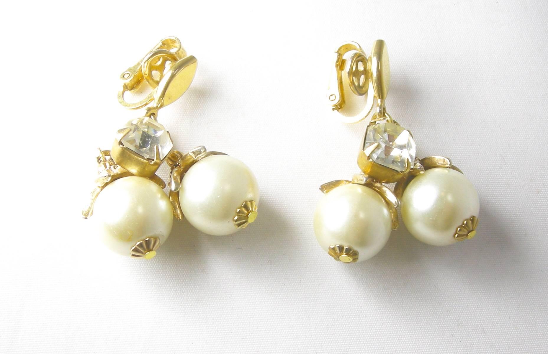 These vintage Trifari clip earrings have two large round faux pearls that hang on a link chain with a large sized bezel crystal.  They are set in a gold tone metal finish, measure 2” x 1”,  are signed with the crown “Trifari” and are in excellent
