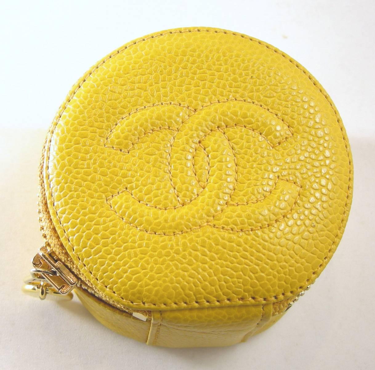 This is an authentic Chanel vintage caviar round jewelry case in mustard yellow.  It is crafted in caviar leather with a Chanel CC logo quilted on the lid. It opens with a wrap-around gold tone  zipper to a black velvet interior. The length 3” and
