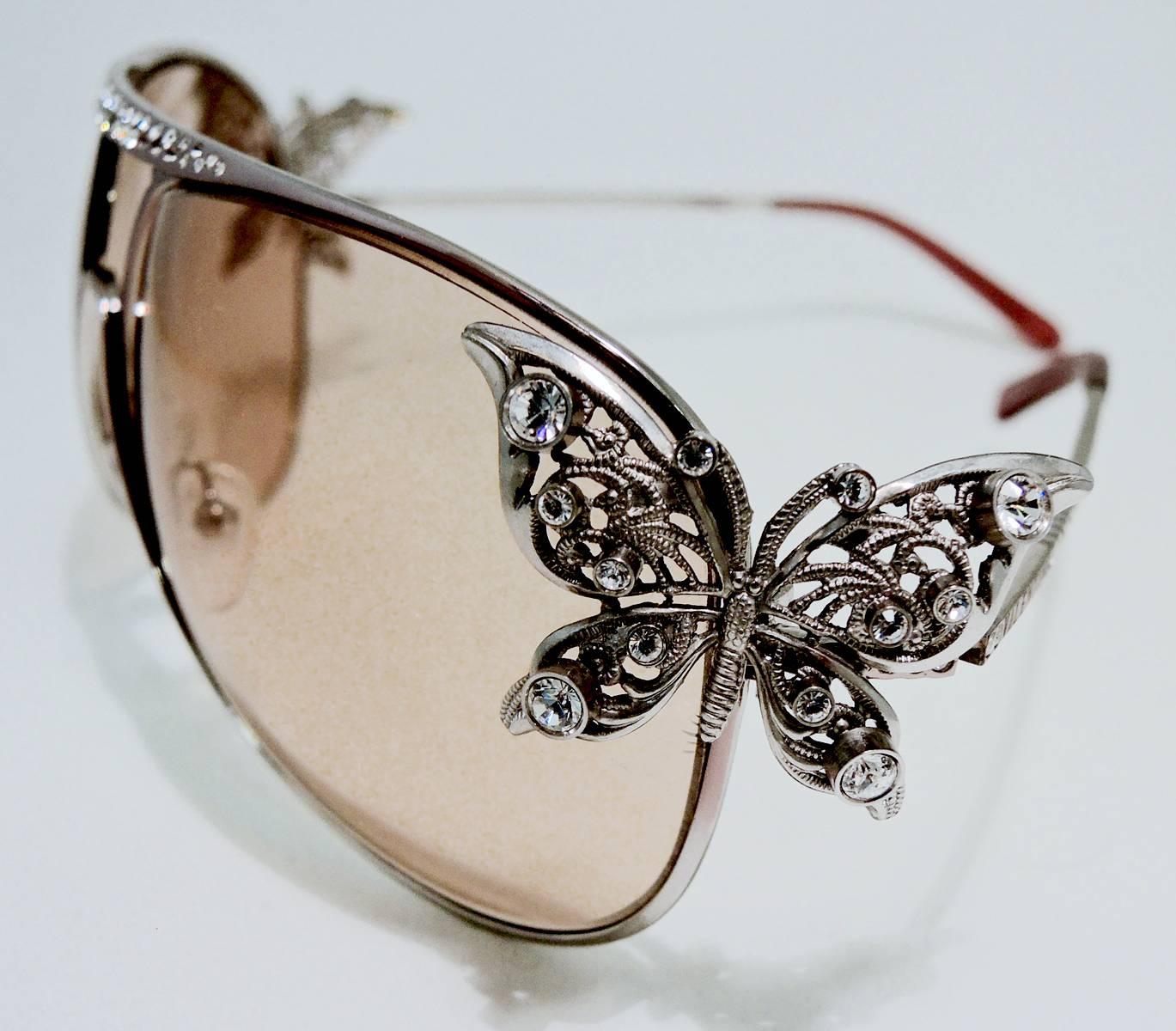 These vintage Valentino sunglasses have their original lenses and have pave crystals in the front. There is a butterfly on each side. They have light pink tint lenses and are a silver tone color. They measure 6” x 2-1/8”, are signed “Valentino” and