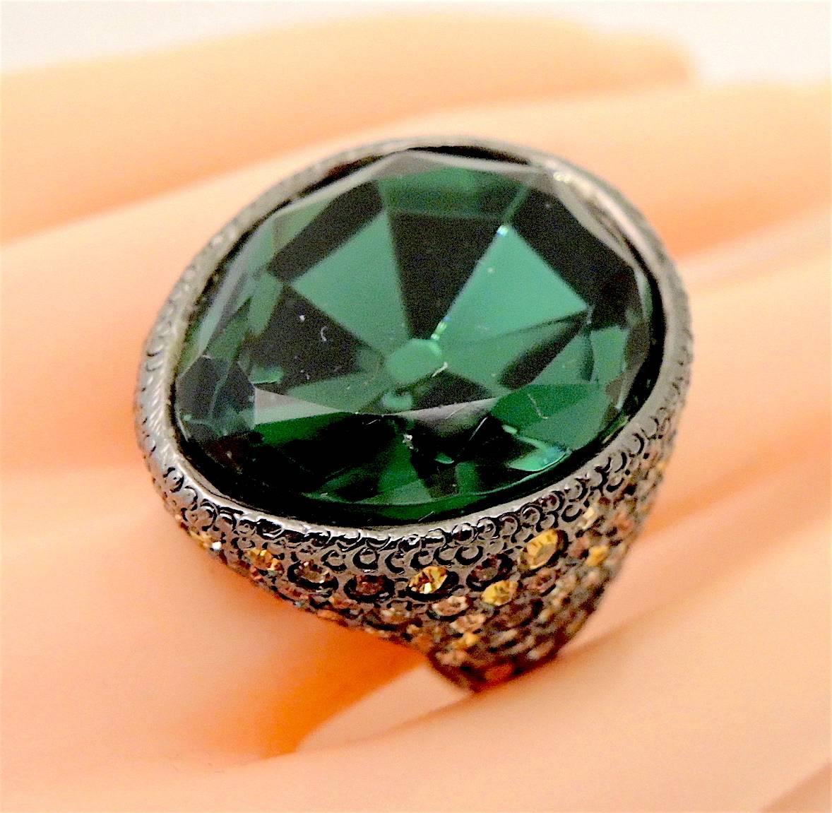 This huge Kenneth Jay Lane ring is a statement maker.  It has a large faceted emerald green crystal center. There are multiple shades of orange crystals on the side. It is set in gunmetal finish and measures 1-1/4” x 1”.  It is adjustable up to a