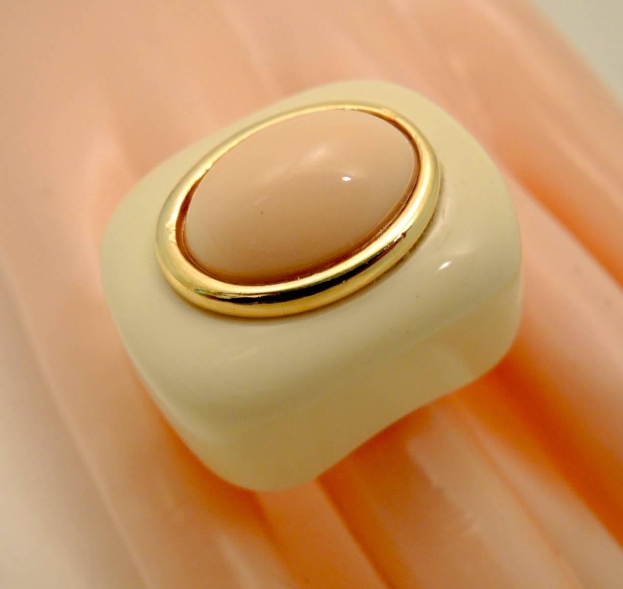 This Kenneth Jay Lane ring features a cream color resin ring with a pink or light coral oval cabochon center stone in a gold-tone bezel frame. The ring measures 1” x 1-1/4” and is adjustable up to a size 7.  It is signed “Kenneth Lane” and is in