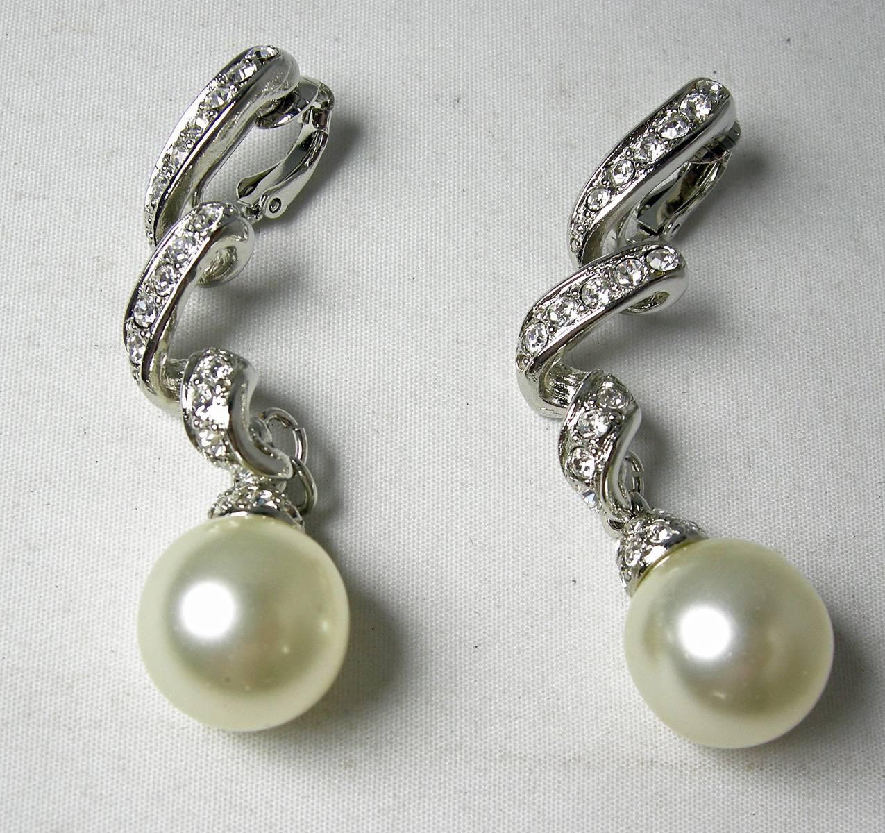 These sparkling clip earrings display gorgeous faux drop pearls that dangle from a swirl design encrusted with rhinestones. The bottom of the earrings has pave rhinestone caps. The earrings are made in a silver tone rhodium setting.  They measure 2”