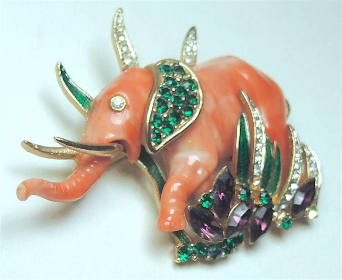 This famous 1950s brooch by Hattie Carnegie features a coral color resin elephant figure with green, clear and purple rhinestone accents in a gold-tone setting. Although unmarked, it is an acknowledged Carnegie design.  It measures 3” x 2” and is in