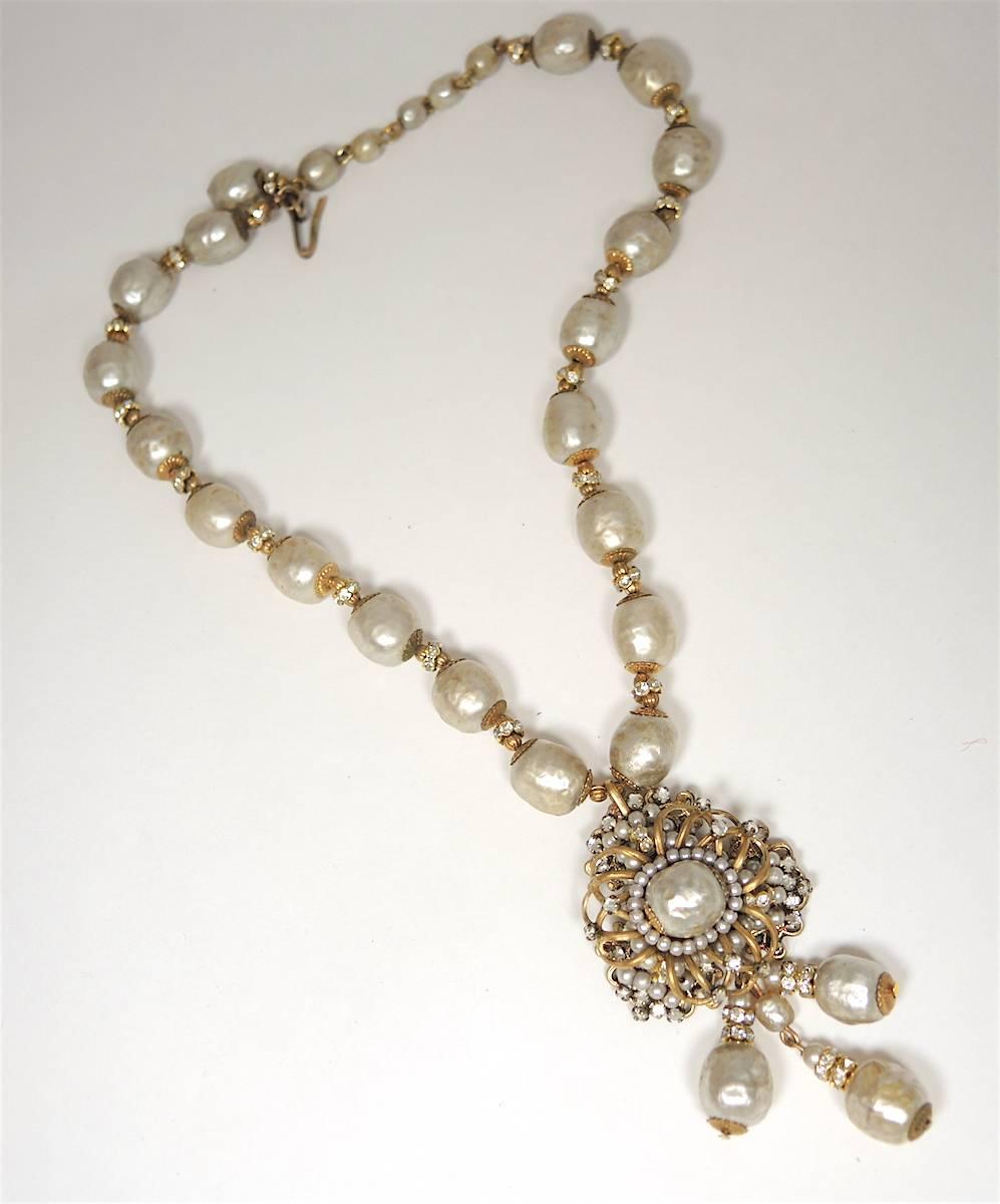 This signed 1950s Miriam Haskell necklace was made with beautiful cream color faux baroque pearls with clear rhinestone accents in a gold tone setting. The centerpiece has a centered oval baroque pearl framed by seed pearls. There are 3 pearl drops