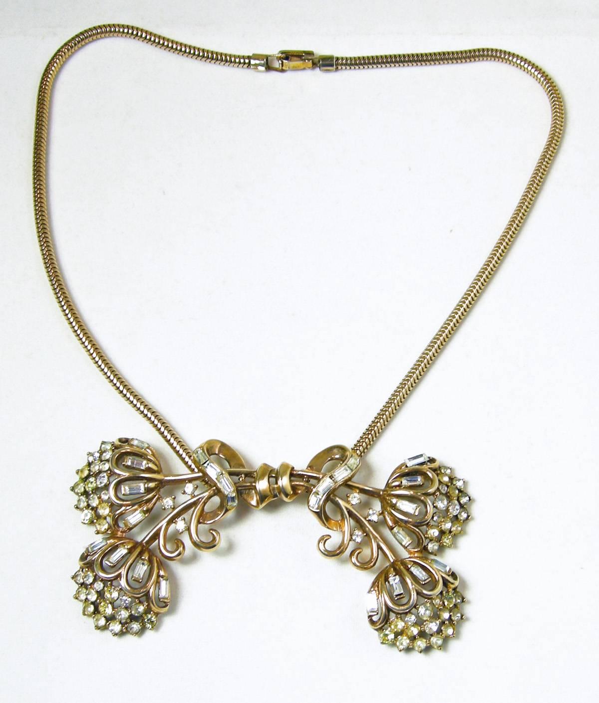 This vintage Trifari necklace is from the 1940s and has an abstract floral bow design accentuated with rhinestones.  It has a gold tone snake chain that is 15” long with a fold over clasp.  The chain leads downward to the centerpiece that measures