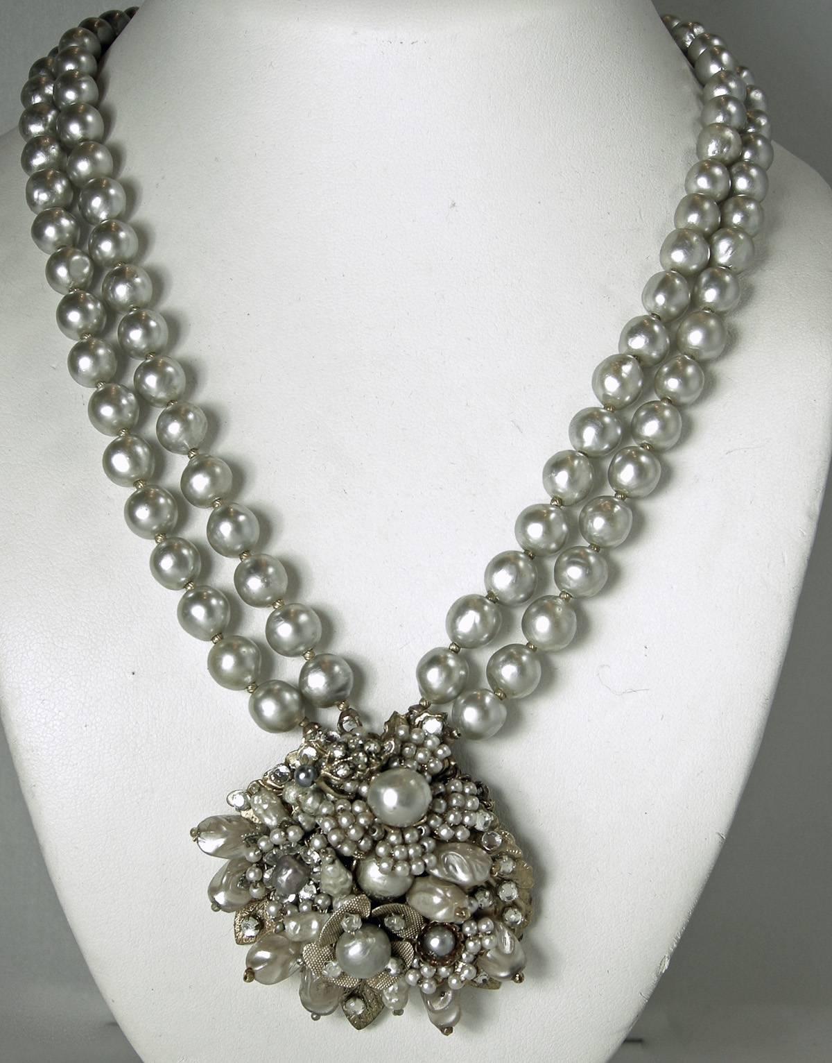 This vintage 1950s double strand Miriam Haskell necklace has faux gray pearls with small golden beads in between. It leads downward toward a centerpiece that has a golden flower at the bottom and three floral clusters of faux seed pearls. The border