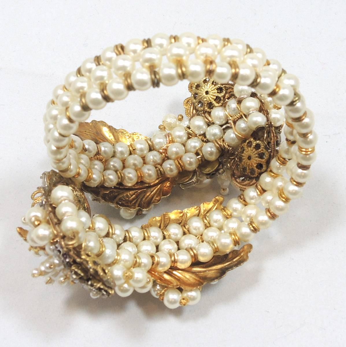 This Miriam Haskell coiled faux pearl bracelet has 3 rows of pave set faux pearls throughout the bracelet. It has a floral design with 10 flowers. All of them have large rhinestones with a pearl in the center. This bracelet is in a gold tone setting