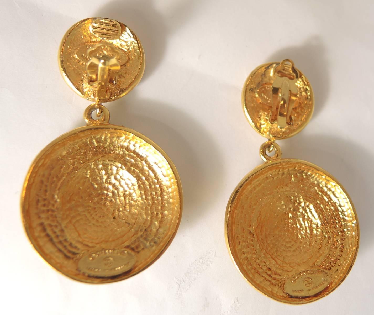 These Chanel round drop disk earrings have a small button top and a larger round disk at the bottom with the monogrammed letter “X” all over them. They are in a gold tone setting and measure 2-1/2” x 1-1/4”. These clip earrings are signed “Chanel”
