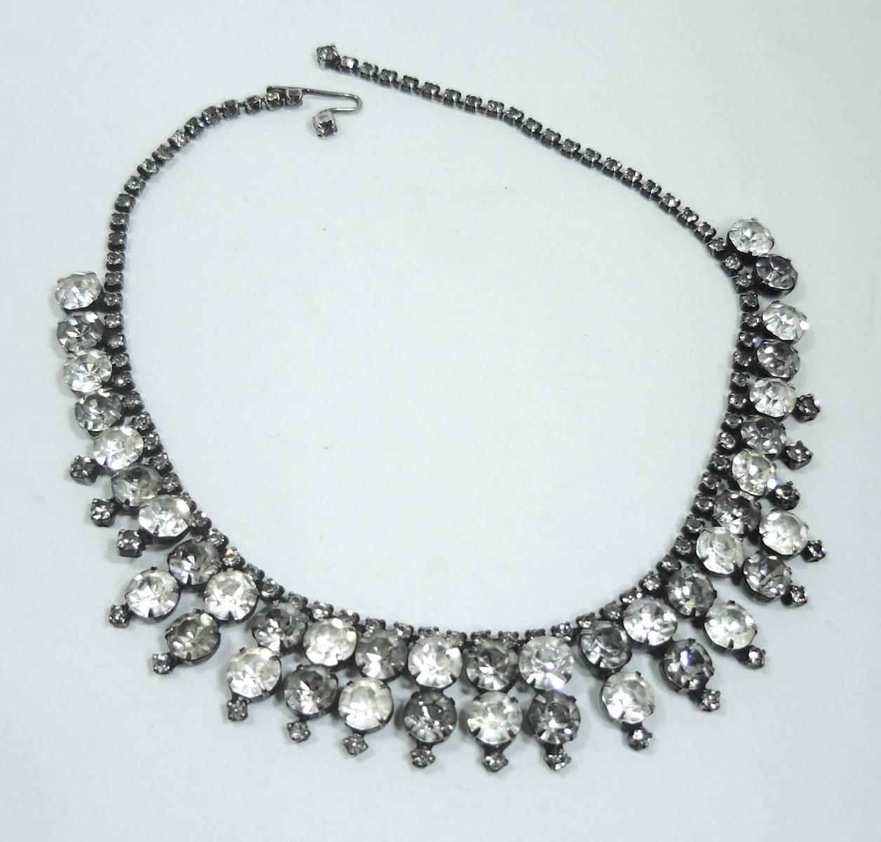 This fabulous necklace was designed by Kramer and has clear rhinestones alternating with a darker grayish rhinestone. The centerpiece has a second row of rhinestones. The second row drapes with smaller rhinestones at the tips. It has a pewter tone