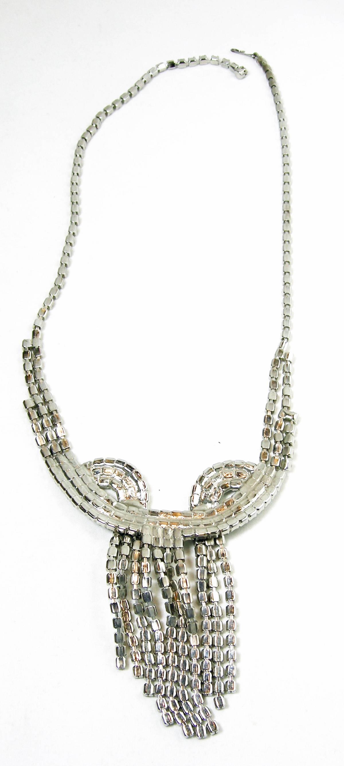 This vintage 1950s rhinestone necklace features a centerpiece that has a looped design with 10 cascading rhinestone strands that form a wide tassel. There are 3 strands on each side but they lead to a single strand necklace. This necklace has a hook