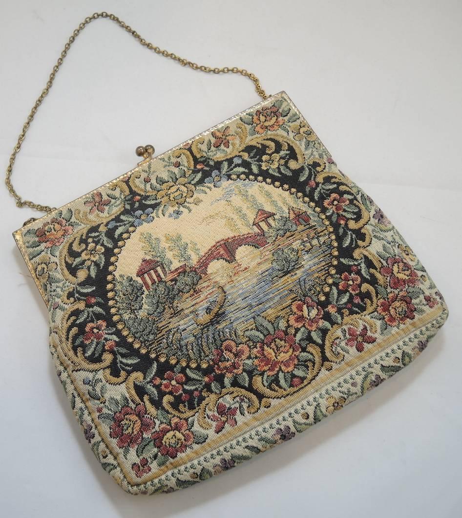 This is a special tapestry purse because it has survived throughout the years in mint condition. It has a picture of water flowing underneath a bridge framed with a floral design and has colors such as pink, beige, maroon and black. It is made with