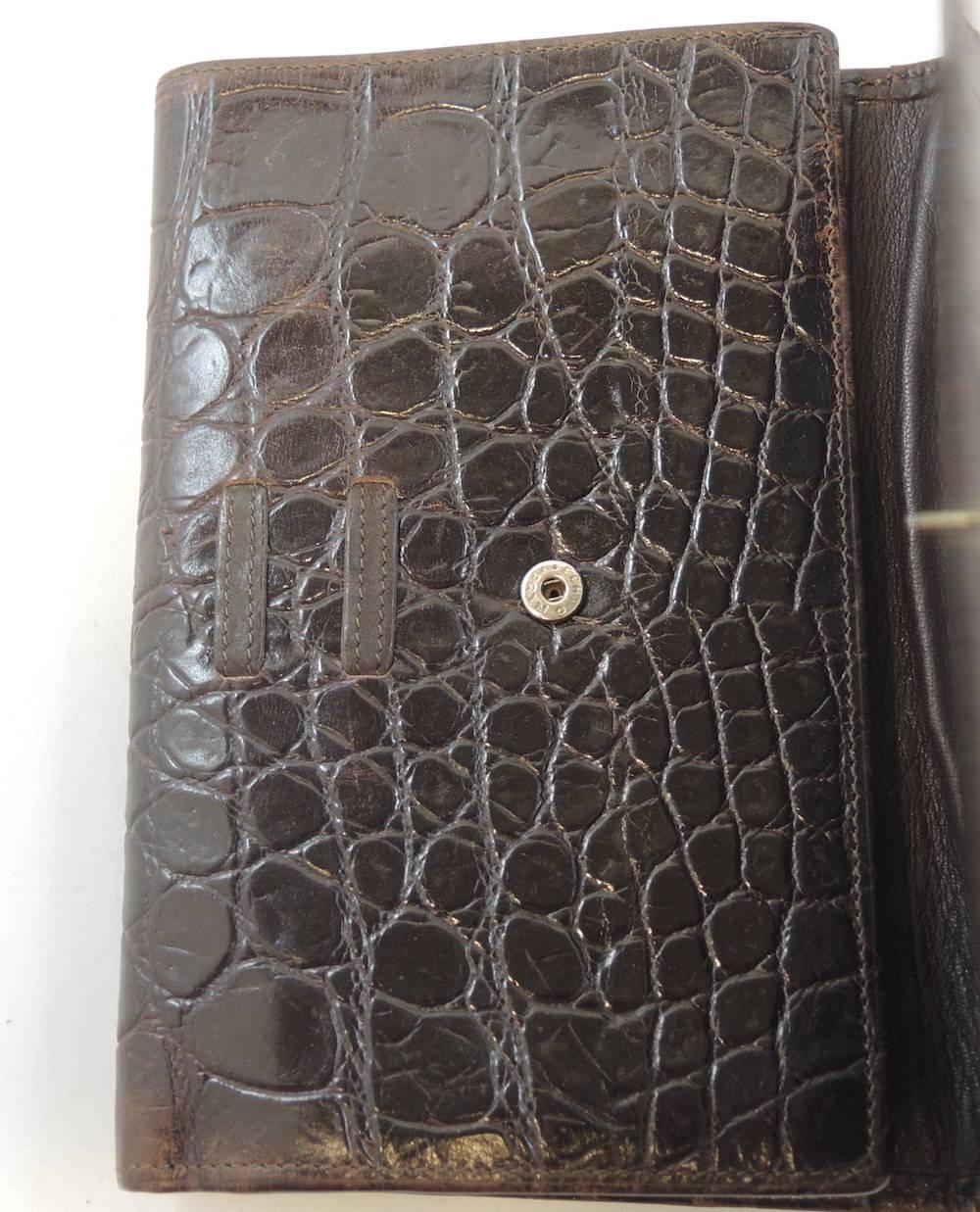 This Gianni Versace crocodile embossed wallet is made of luscious brown leather. The flap has a bow in the front and a snap in closure. The inside opens up to a tri fold style. It has two large bill slots, a coin pocket and room for credit cards. It