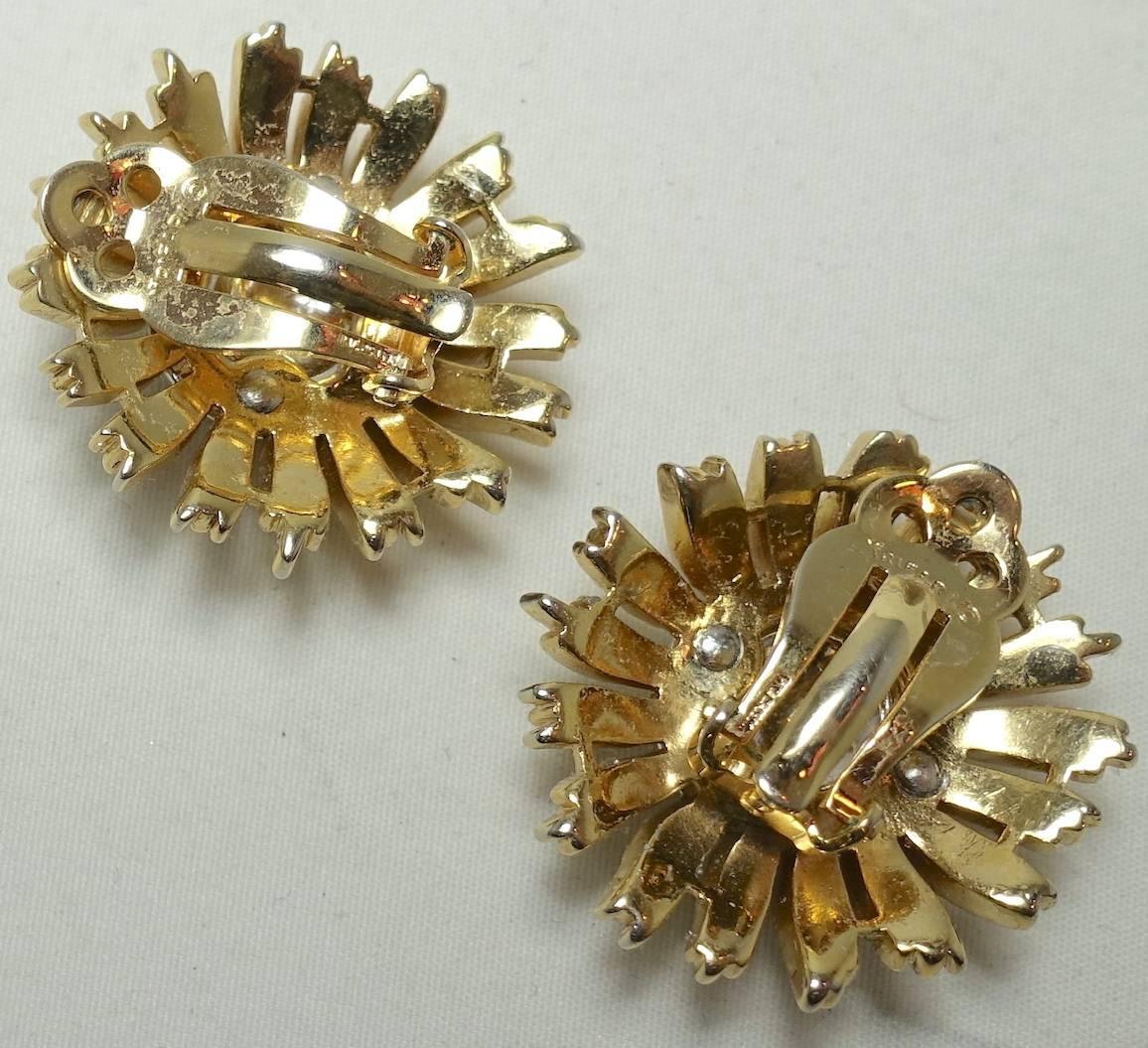 These Trifari 1950s earrings feature clear rhinestones in the center in a floral design. They are in a gold tone setting and measure 1-1/4” x 1-1/4” and are signed “Trifari”. They are in excellent condition.