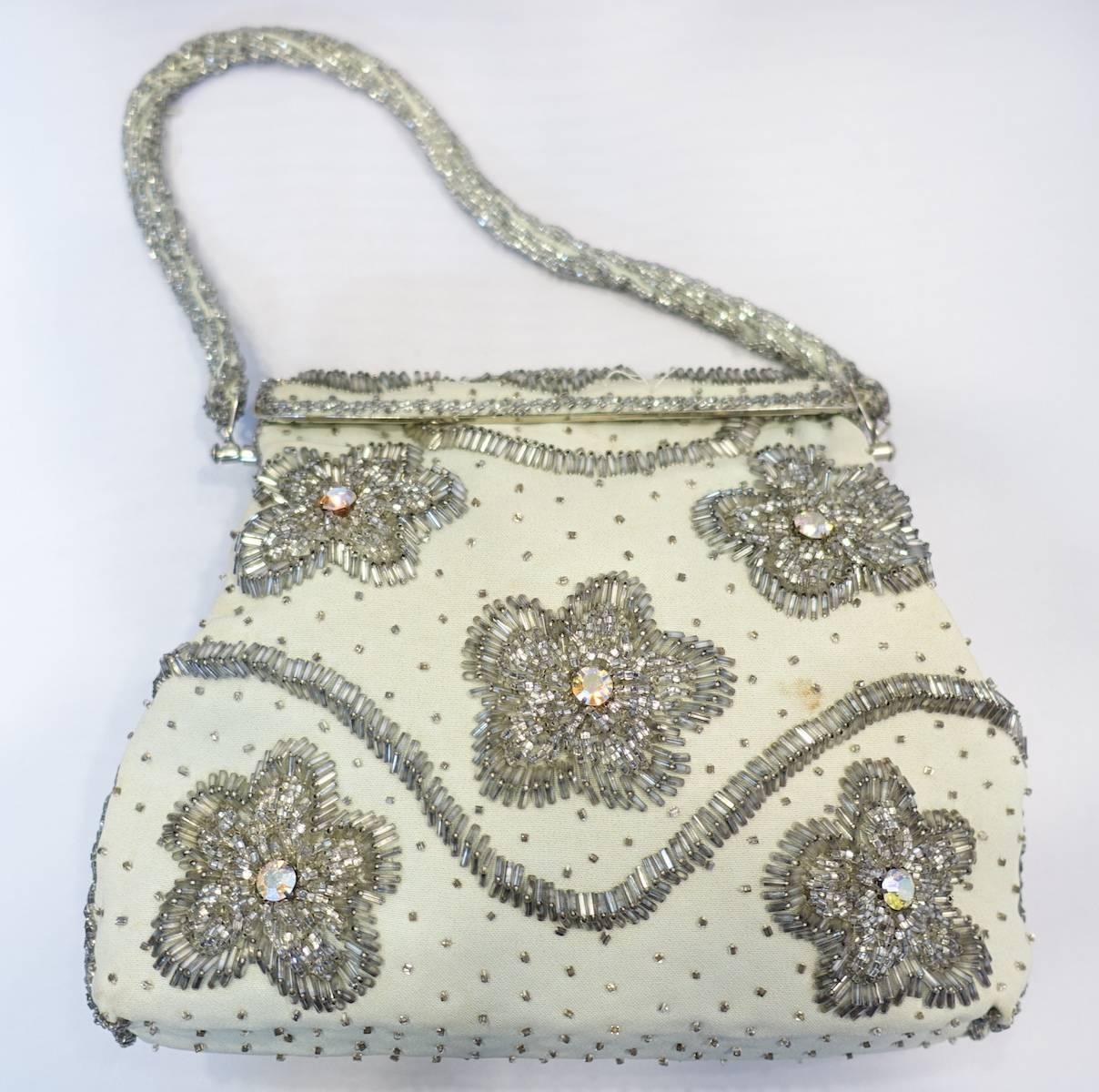 This beautiful1960s hand bag is handmade and consists of gray & clear beads with aurora borealis stone accents on crème color cotton.  This handbag measures 7” high x 8-1/2” wide with a fold-over closure. The beaded strap is 5.5” up from center. 
