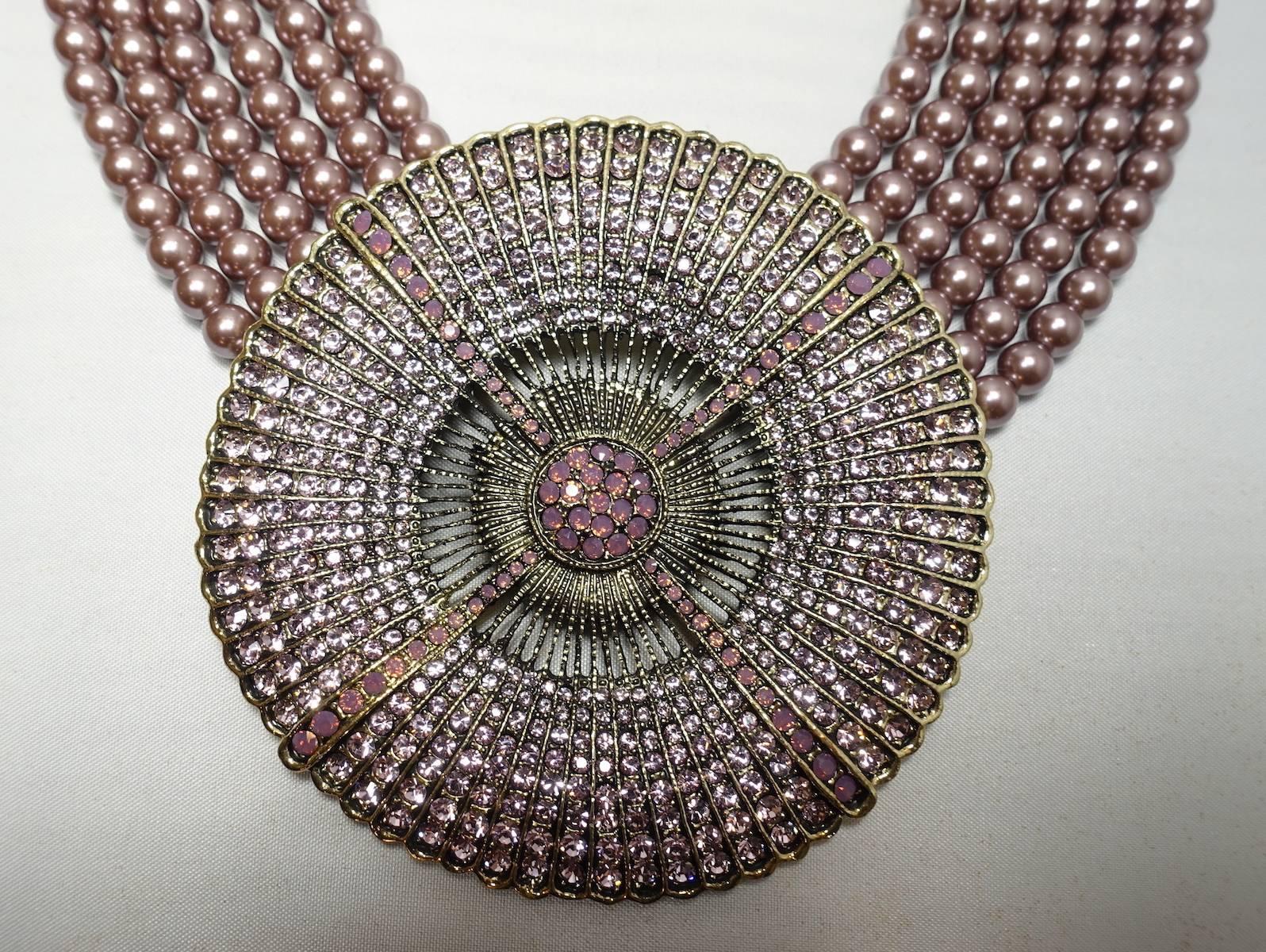 This Heidi Daus Belgium disc drop necklace features 6 rows of mauve color faux pearls. It has a large disc drop that is encrusted with rows of pave set round crystals. It has a scalloped border and is in a bronze tone setting. It measures 16-1/4” X