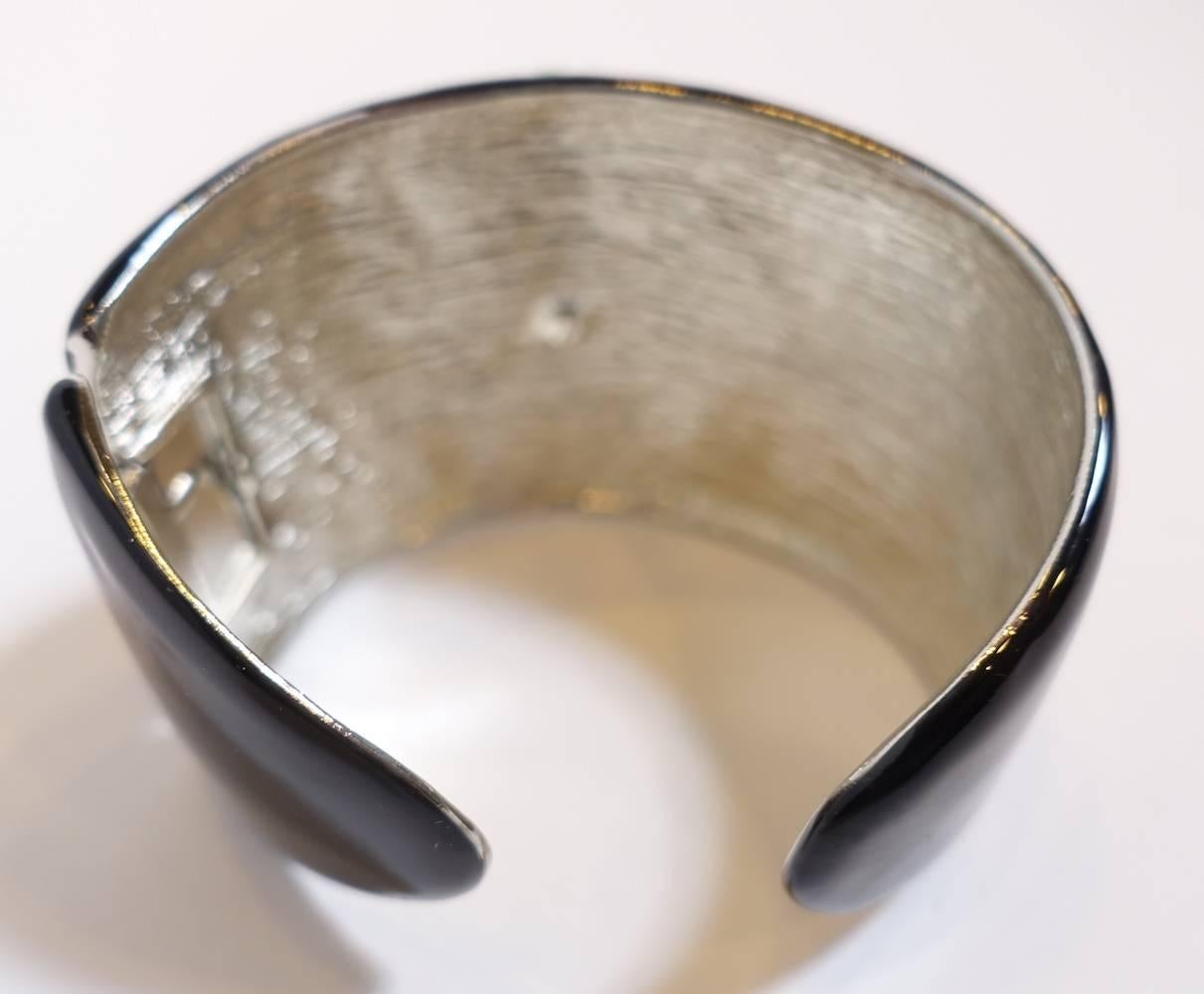 This signed Kenneth Jay Lane deco style cuff features a heavily carved faux jade center with crystal accents on each side on black enamel.  It is in a nickel-free silver-plated base metal setting.  This cuff measures 7” x 1-7/8” wide with a