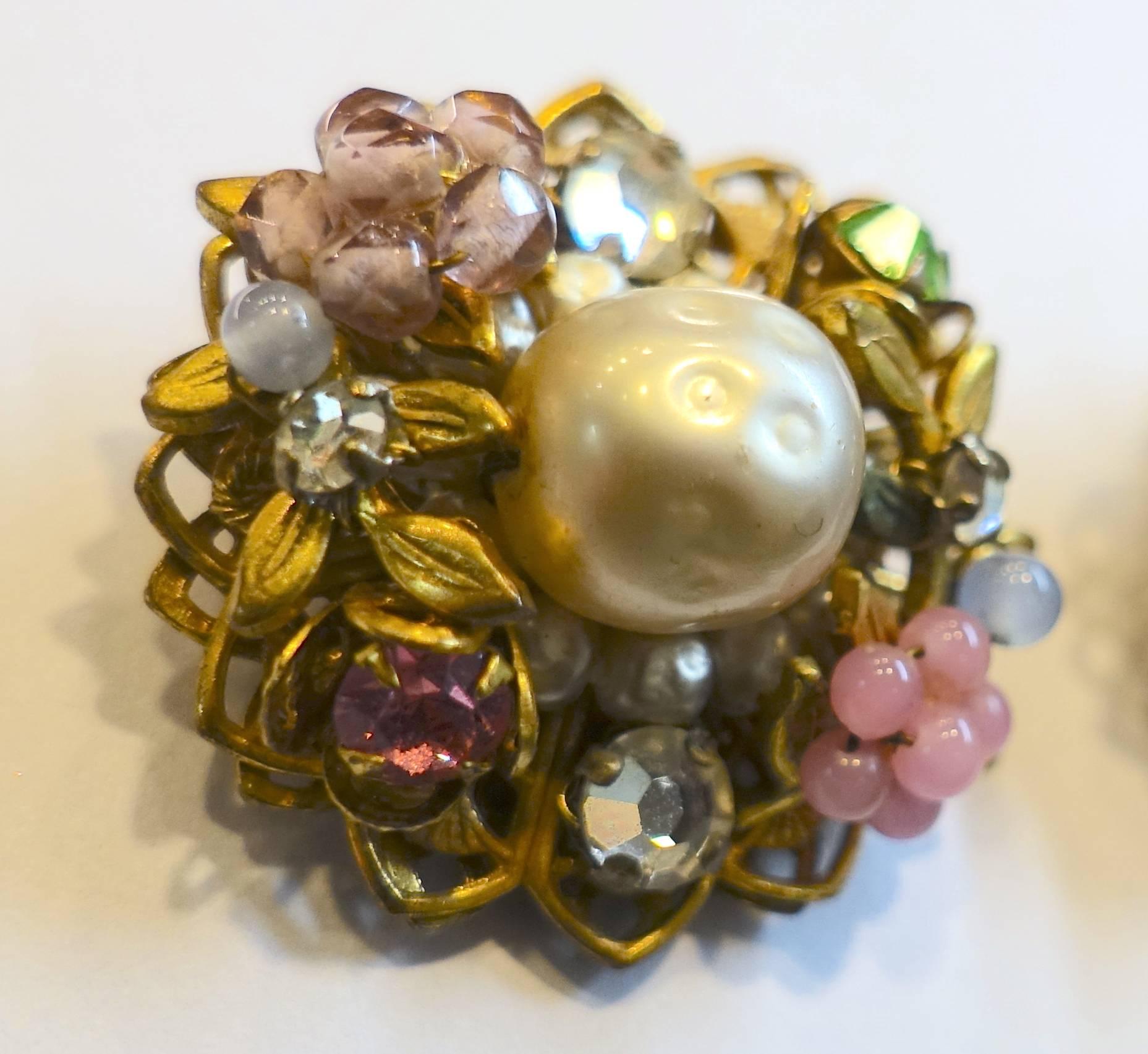 These clip earrings have a large faux baroque pearl center combining lavender bugle beads, blue rhinestones, pink beads that form a flower and green rhinestones lying on gold tone leaves.  It measures 1” and signed “DeMario”.  These earrings are in