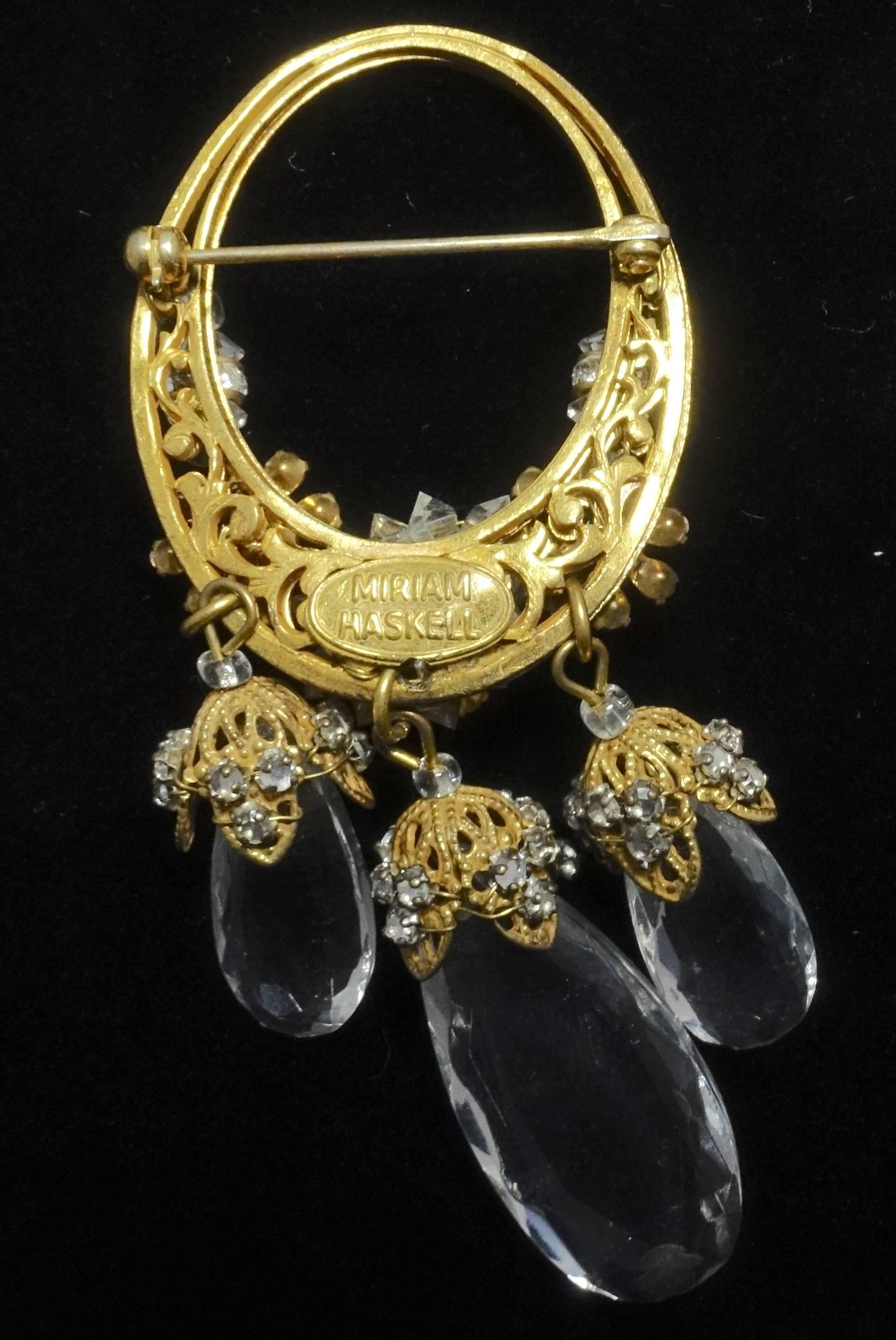 This is a wonderful brooch by Miriam Haskell.  It has a gold tone double oval frame with crystal flowers at the bottom.  There are two small-capped crystals hanging at the bottom with a larger capped crystal in the middle.  It measures 3-3/8” long