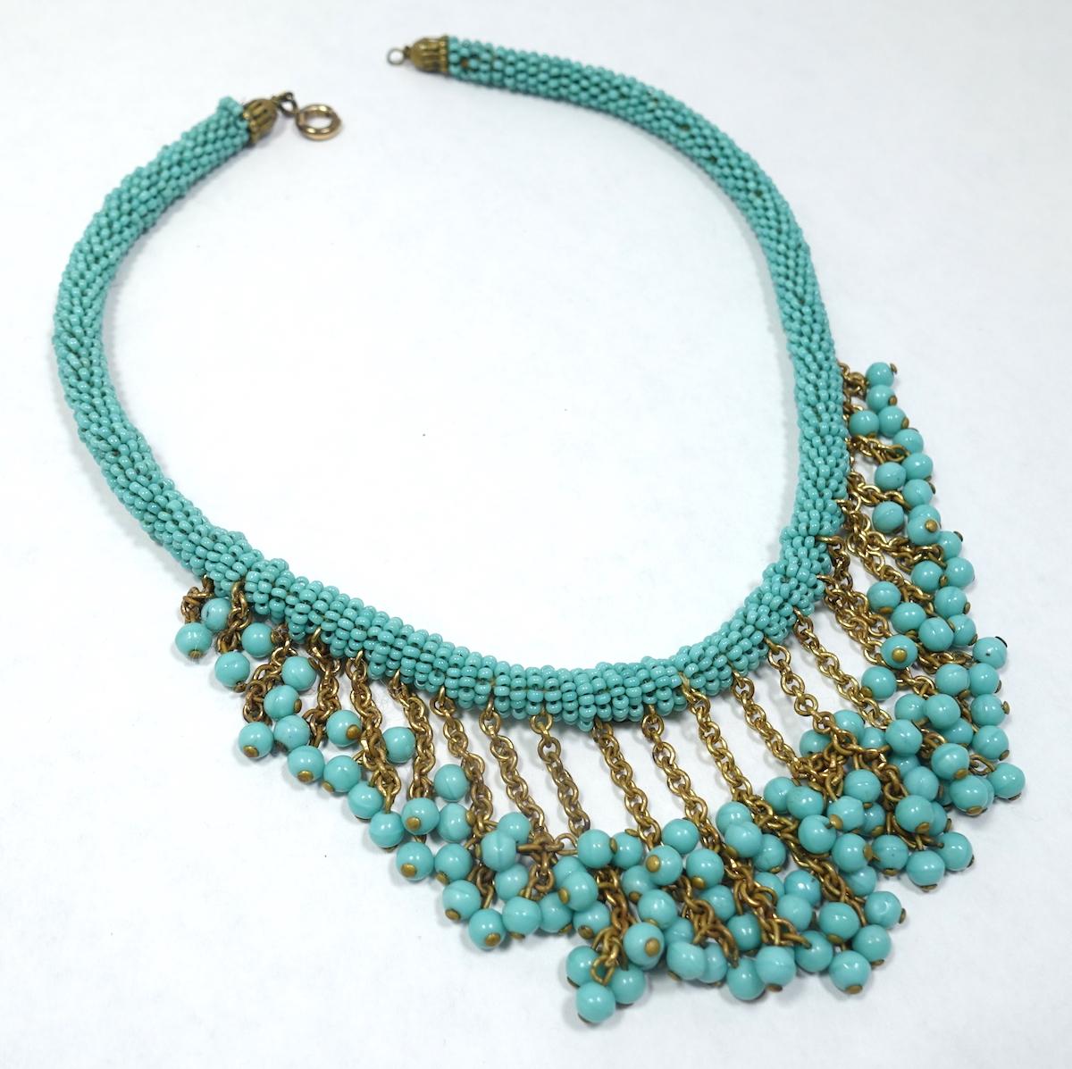 This early vintage 1930s Miriam Haskell necklace features turquoise glass beads going around the neck. Hanging downward are gold tone chains with clusters of glass turquoise beads forming a bib effect. This very early Haskell necklace is in mint