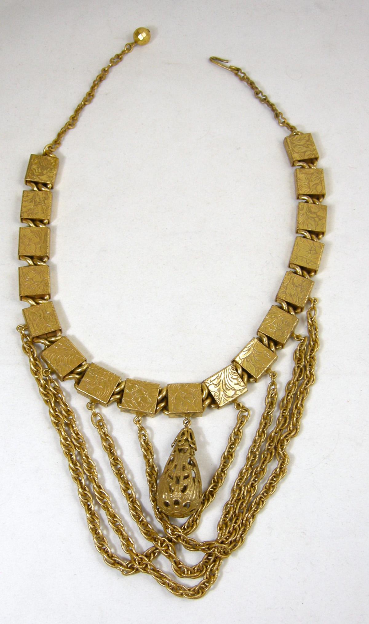 This unique retro bib necklace has ornate engraved boxed links that hold four swirling chains with an ornate gold tone filigree drop.  The necklace is 17-1/2” long with a 6” wide with q chain center.  The chains drop approx 4” and the filigree drop