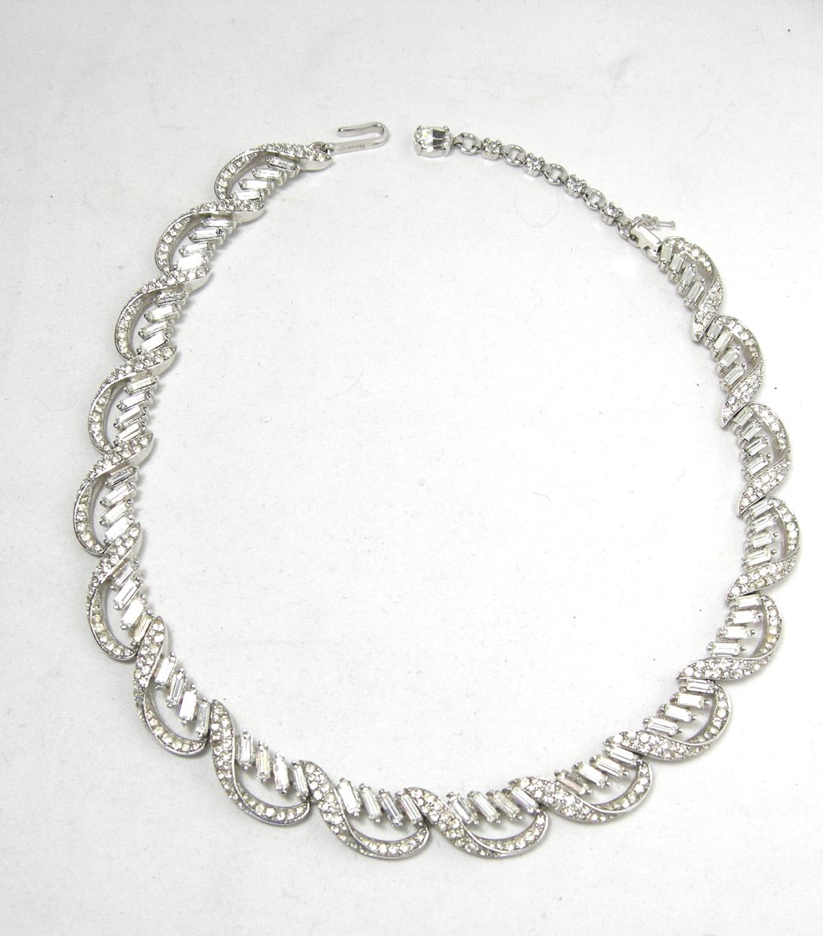 This signed Trifari has scalloped baguette rhinestones going around the necklace.  It is 15-1/2” x 1/2” wide with a hook clasp.  It has silver tone rhodium finish and is signed “Trifari”.   It also has the “T” hangtag and is in excellent condition.