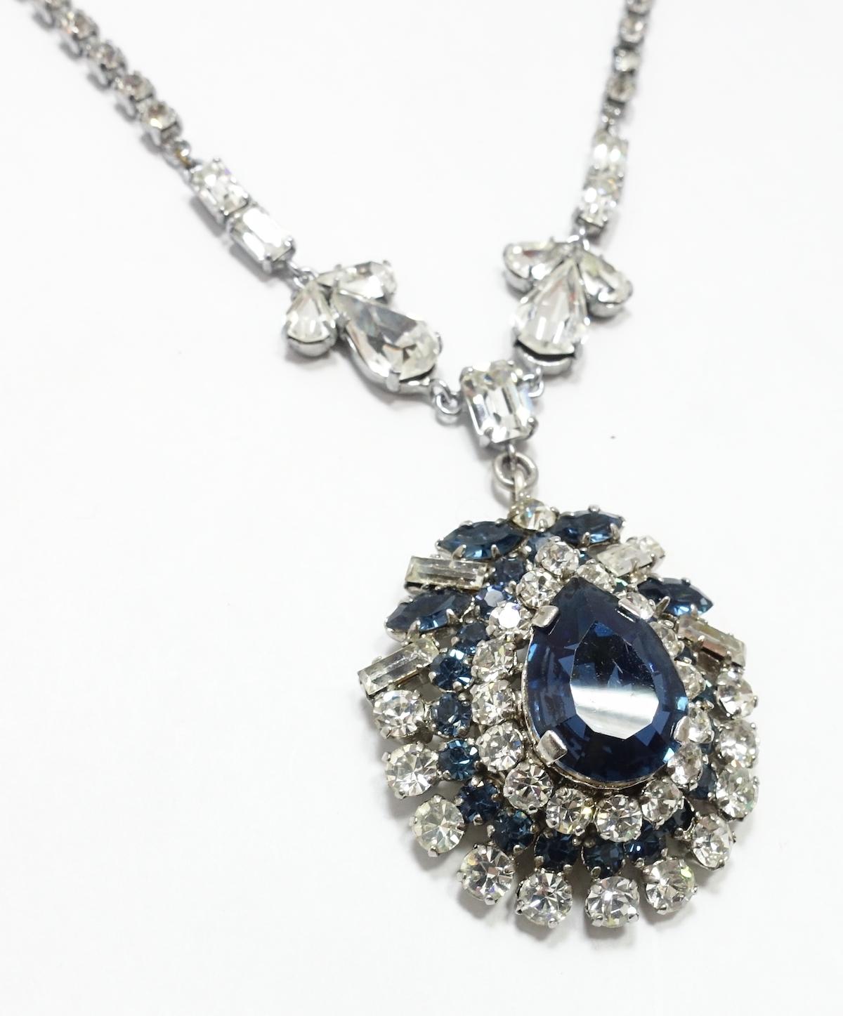This vintage necklace features a teardrop pendant with blue and clear crystals in a silver-tone metal setting.  In excellent condition, the necklace measures 14” x 1/8” with a spring closure; the pendant is 1-1/2” x 1-3/8”. It is an unsigned piece,