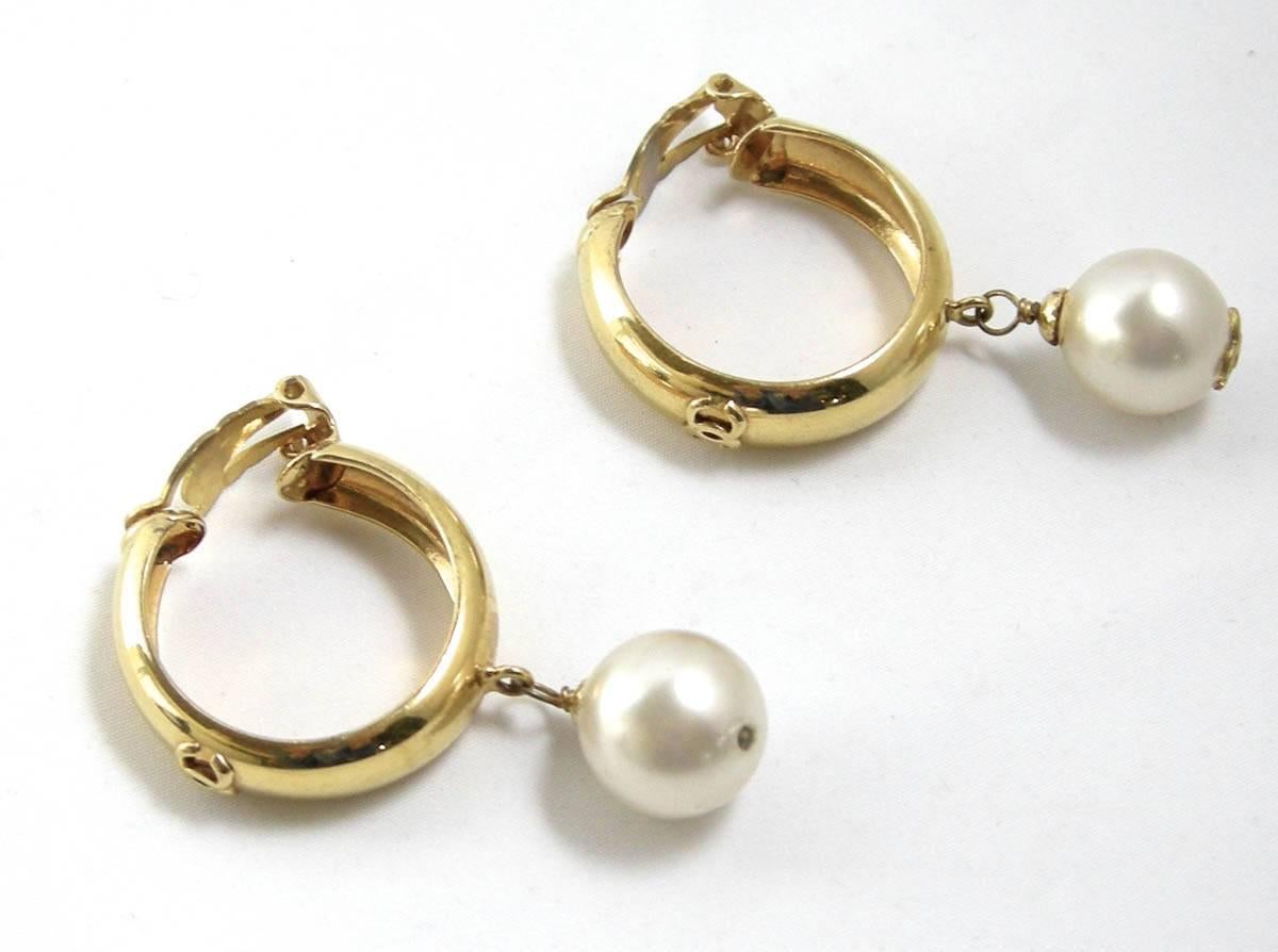 These vintage Chanel clip on earrings feature a hoop in a high polished gold tone metal setting with a faux large pearl drop hanging from the golden hoop. It has the cc hallmark at the bottom of one of the pearl drops. These earrings are from season