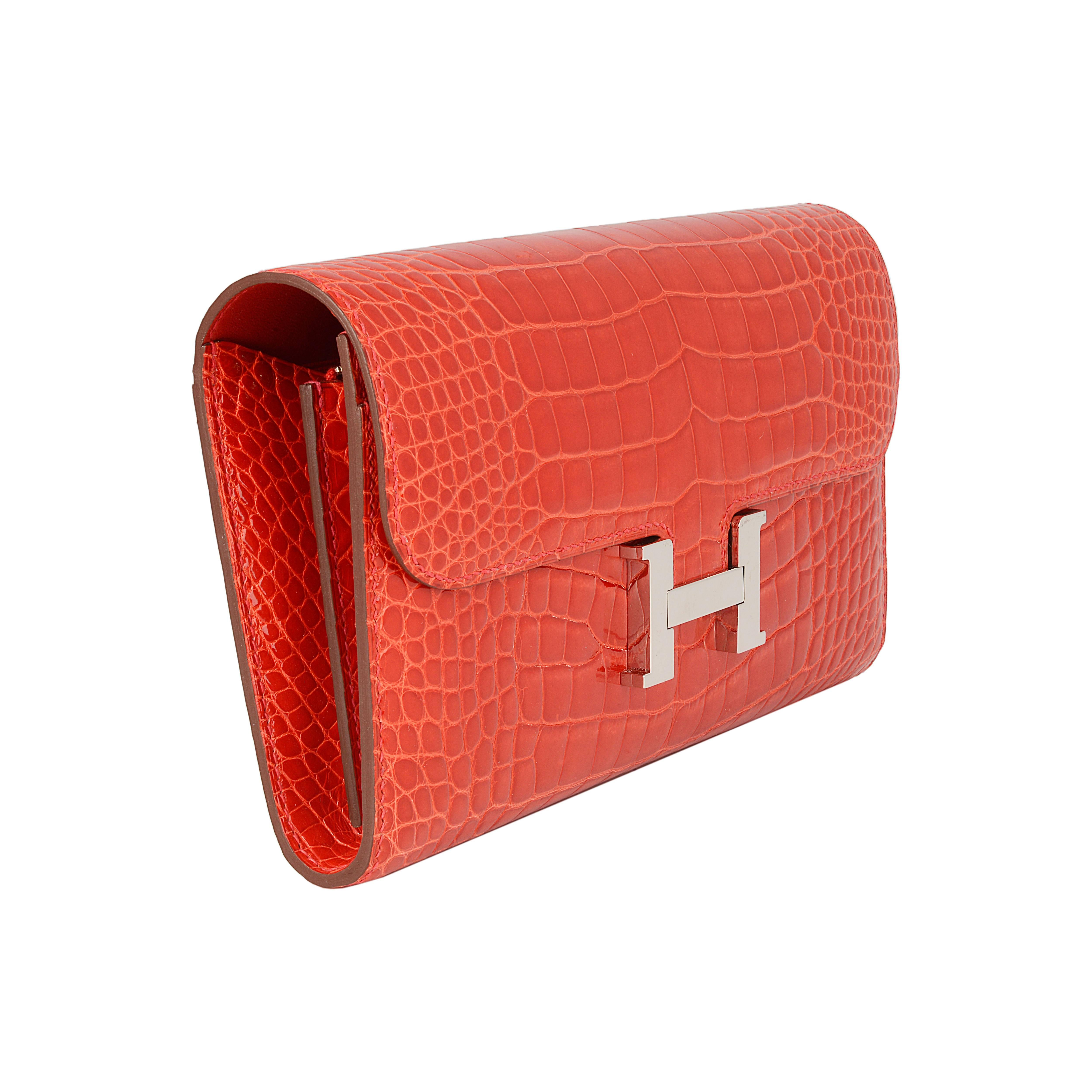 This Hermès Constance Long Wallet, which can also be worn as a clutch bag, is primarily made from red shiny alligator leather accentuated by palladium hardware and the iconic H clip. The interior is lined with red Epsom leather, has two front wall
