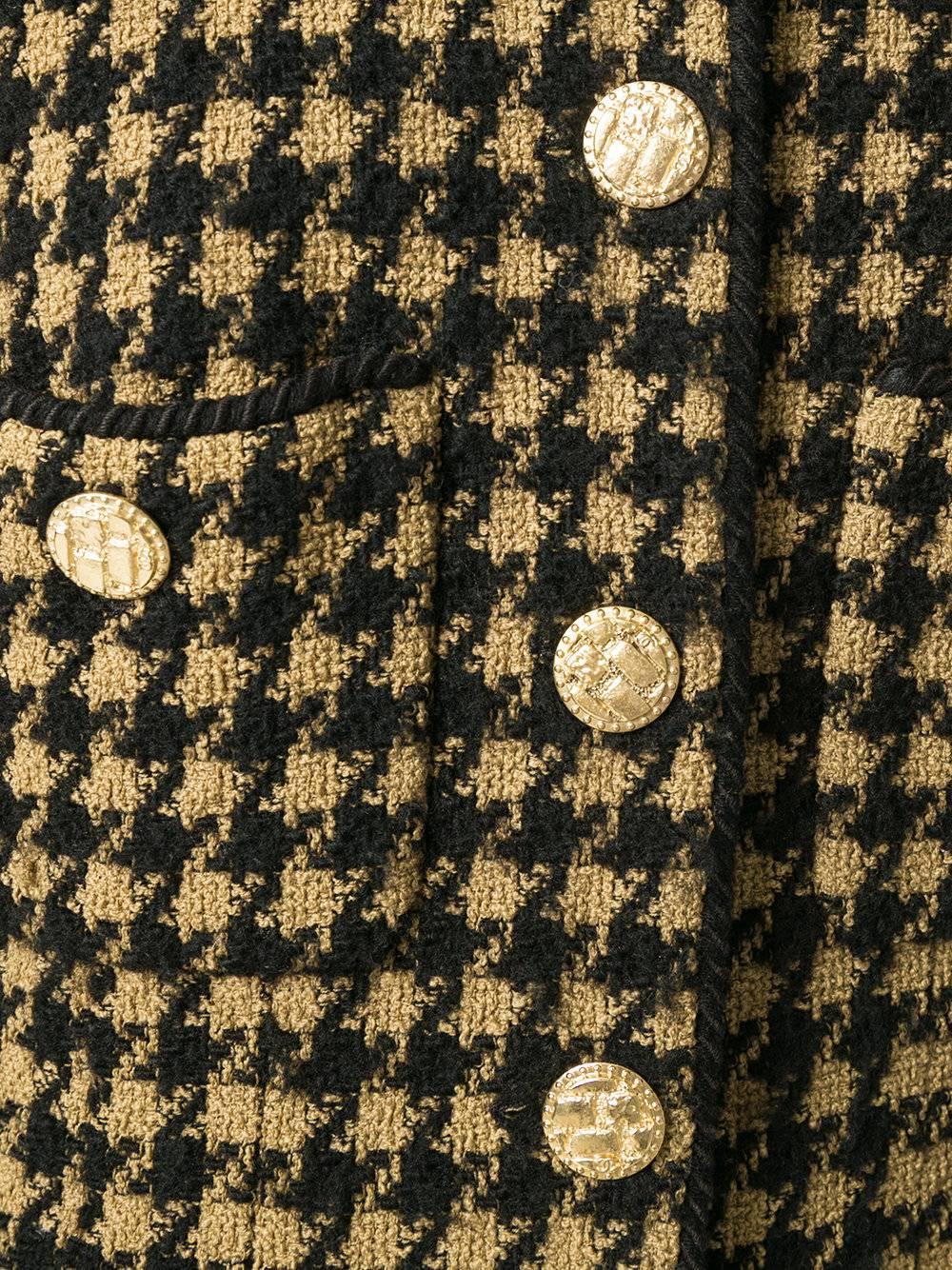 This wool jacket is in a classic camel and black houndstooth pattern. Collarless, the neck is fastened by embossed gold-tone buttons. Featuring four frontal pockets, also embellished by buttons, as are the cuffs. 

Colour: Camel & Black

Material: