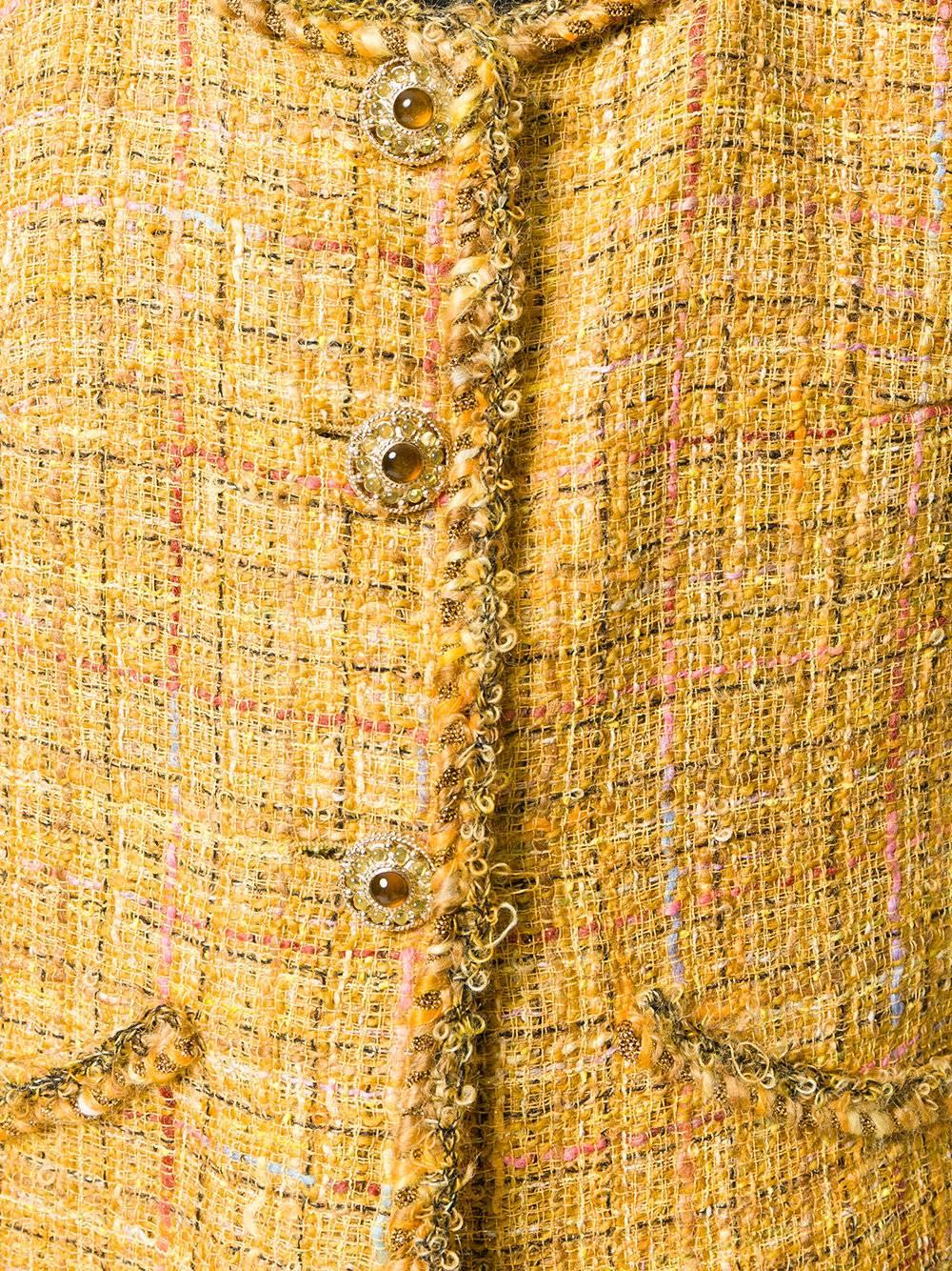 This vintage Chanel twinset is woven from tweed in a deep saffron hue, underscored by threads toned in sparkly gold, pastels and black. Its straight-fitted waistcoat fastens with gold-tone medallions inlaid with gems. The mini skirt is cleaved at
