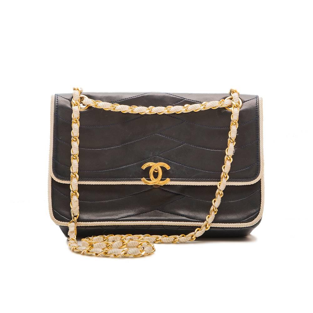 This Chanel Vintage navy leather bag features cream rope piping around the edges which continues throughout the woven gold and cream shoulder chain. The iconic logo fastening opens to reveal one zip pocket and two open pockets on the inside of the