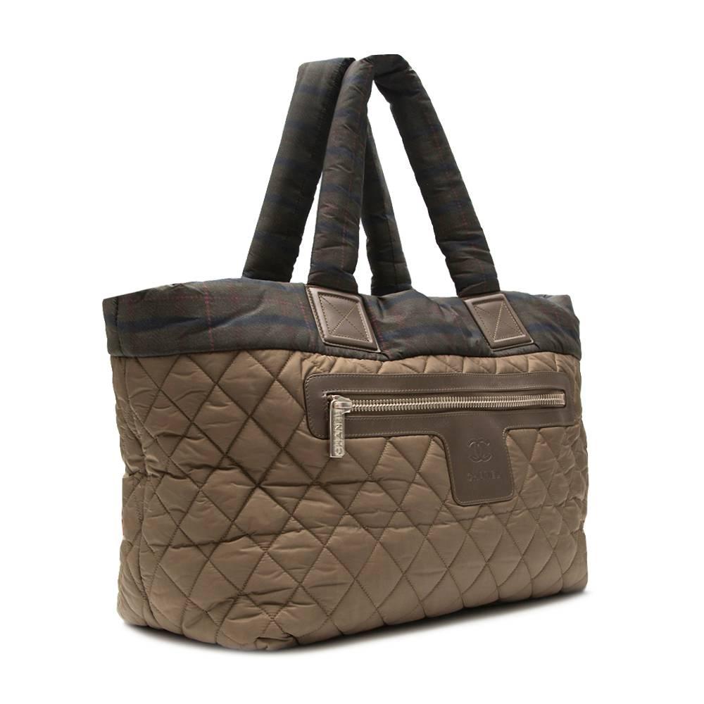This Chanel Coco Cocoon tote bag showcases a spacious, lightweight khaki fabric with a Scottish flair. It opens to a roomy interior with side zip and FOB for your keys, perfect for daily essentials. A stand-out, chic tote made for light travels.