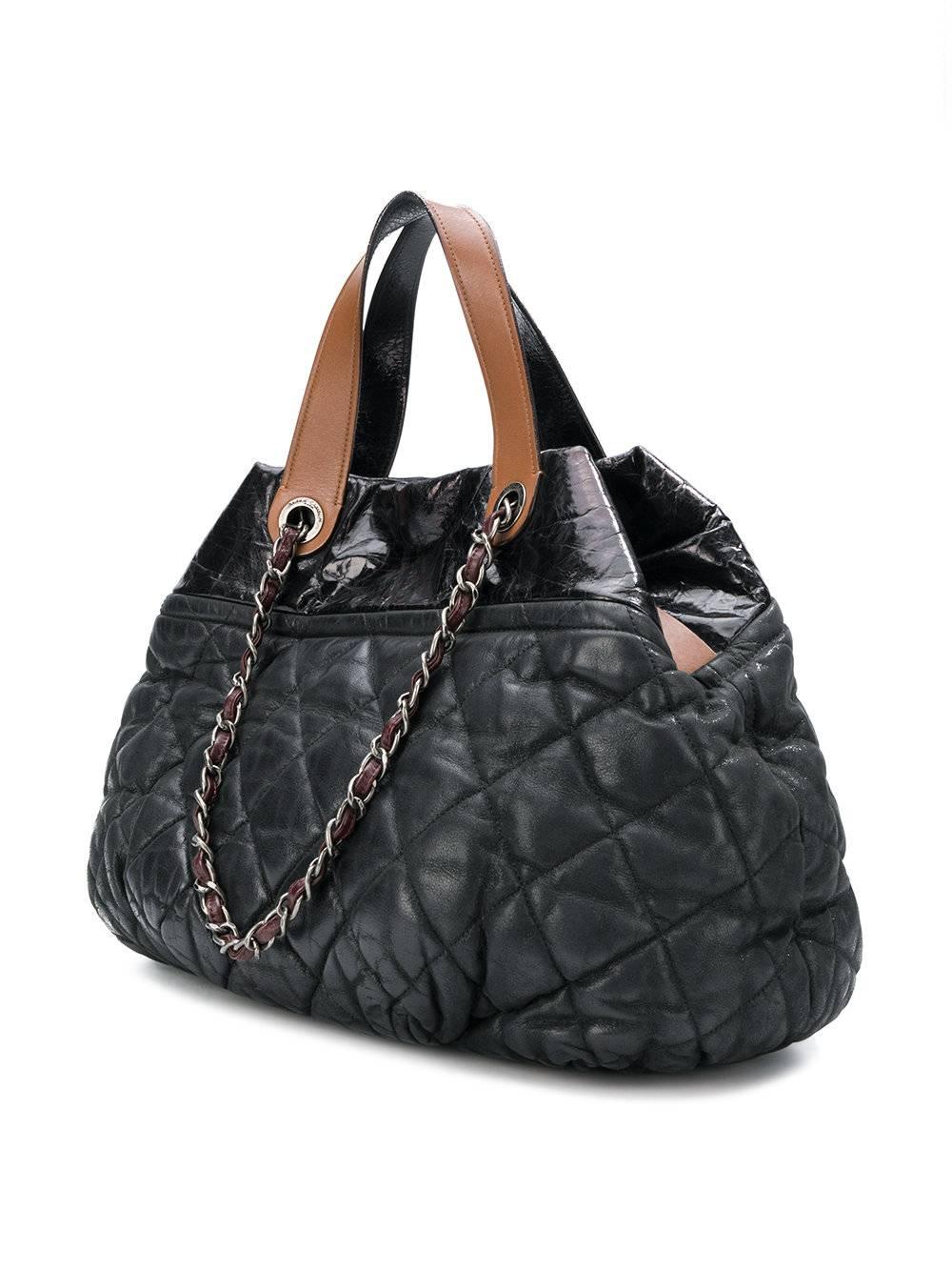 This Chanel Coco Cocoon tote bag showcases a spacious, lightweight gunmetal grey fabric with a quilted finish. It opens to reveal a roomy burgundy interior with two open slip pockets, one larger zipped pocket and fob for your keys, perfect for all