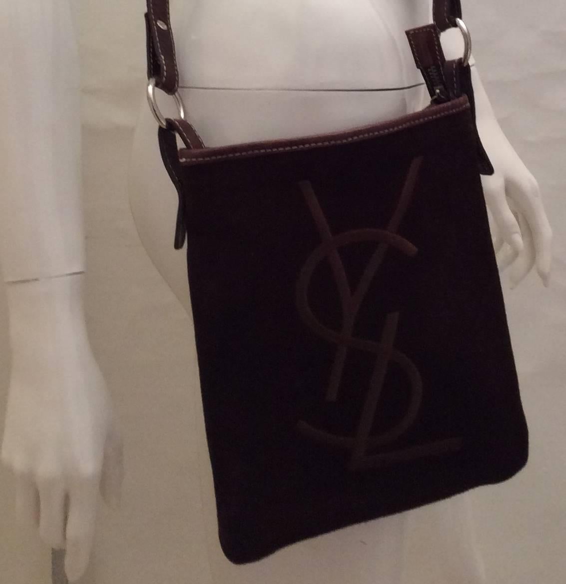 Presented here is an Yves Saint Laurent cross body bag. The bag is brown suede with smooth brown leather trim. There is a large embroidered 'YSL' emblem on the front of the bag. There is white stitching on the bag. The interior lining is an animal