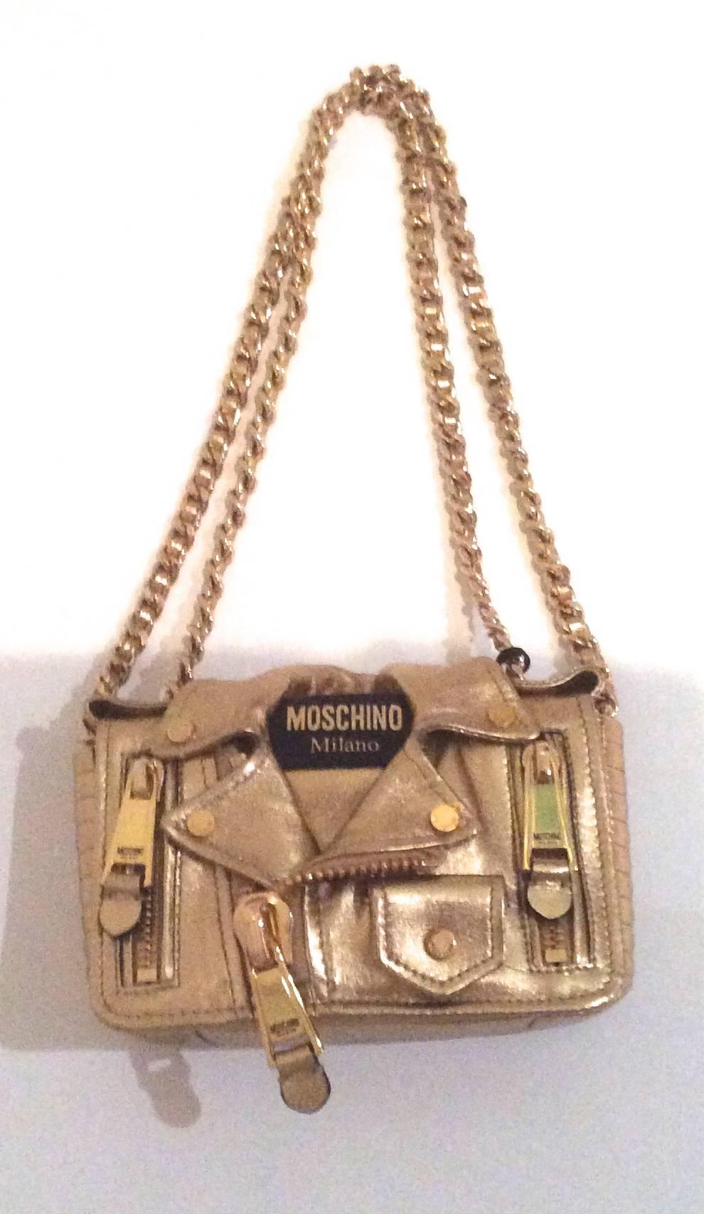 This gold biker Moschino handbag is designed to resemble a gold toned shiny leather jacket. The front of the bag has large zippers with small pockets on the front of the bag. The bag has a snap closure and the cover of the front of the jacket lifts