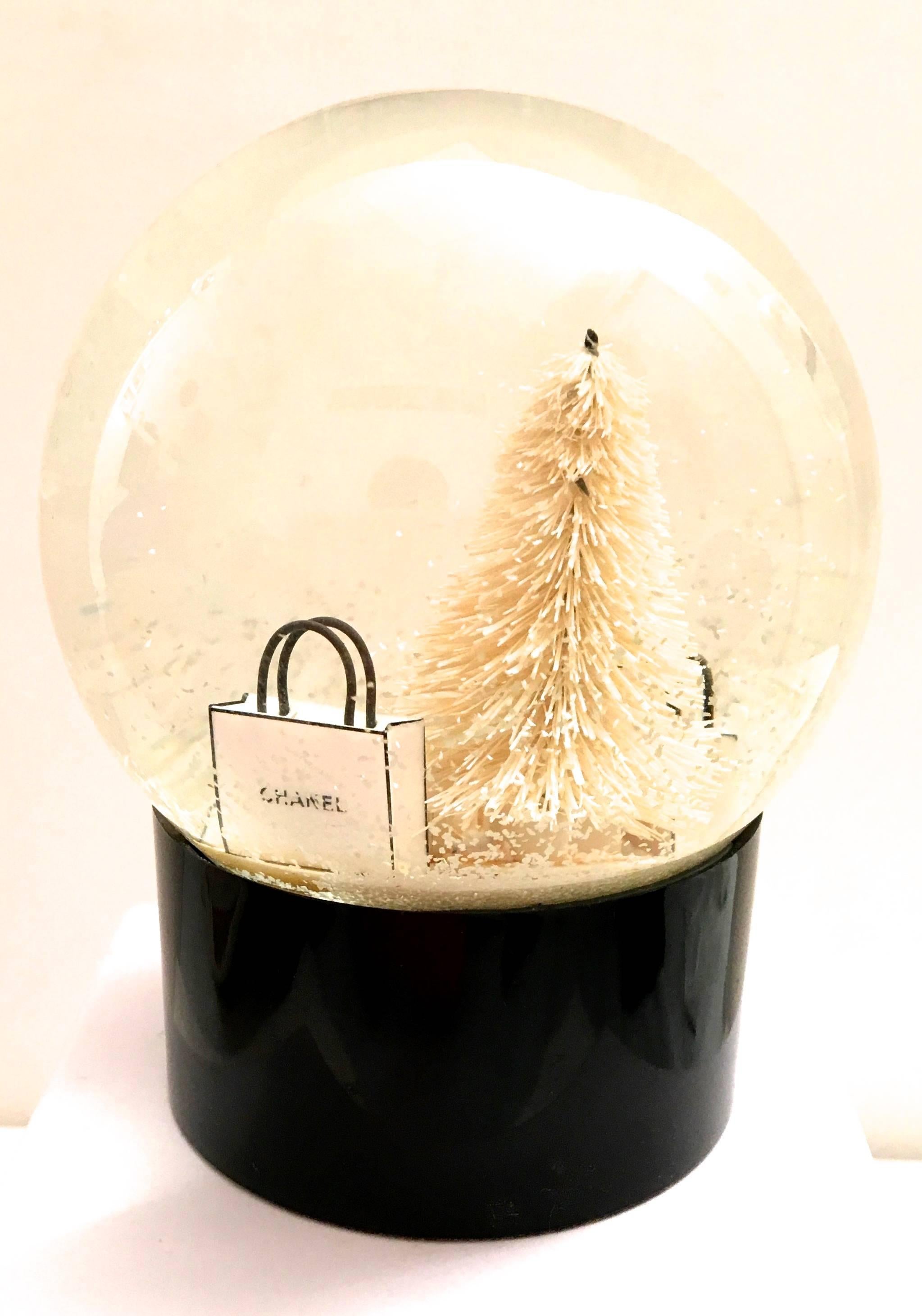 Presented here is a rare Chanel snow globe. The globe is comprised of a glass snow globe set atop a black base that is signed 'Chanel.' The snow globe has a white tree inside the glass surrounded by packages and bags that are signed 'Chanel.'    The
