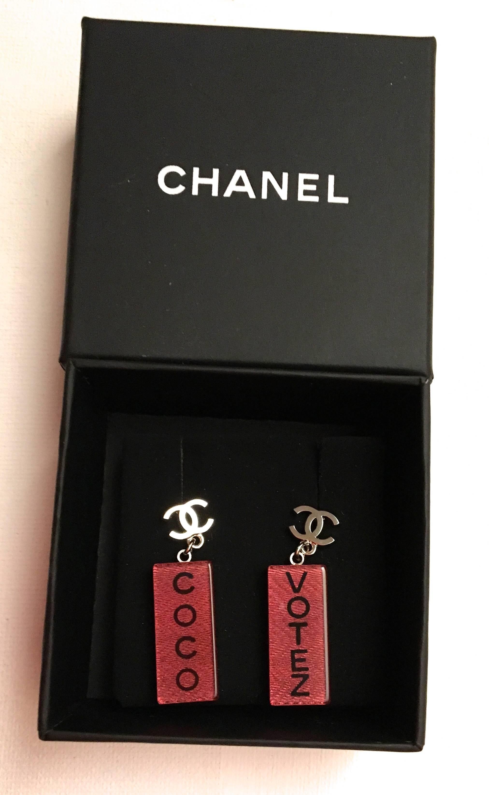 Presented here is a new pair of Chanel lucite earrings. Each earring has a silver tone CC logo with dangling lucite rectangles with a pink / red color tone on either side of the earring. One earring says 'Votez' and the other says 'Coco.' The