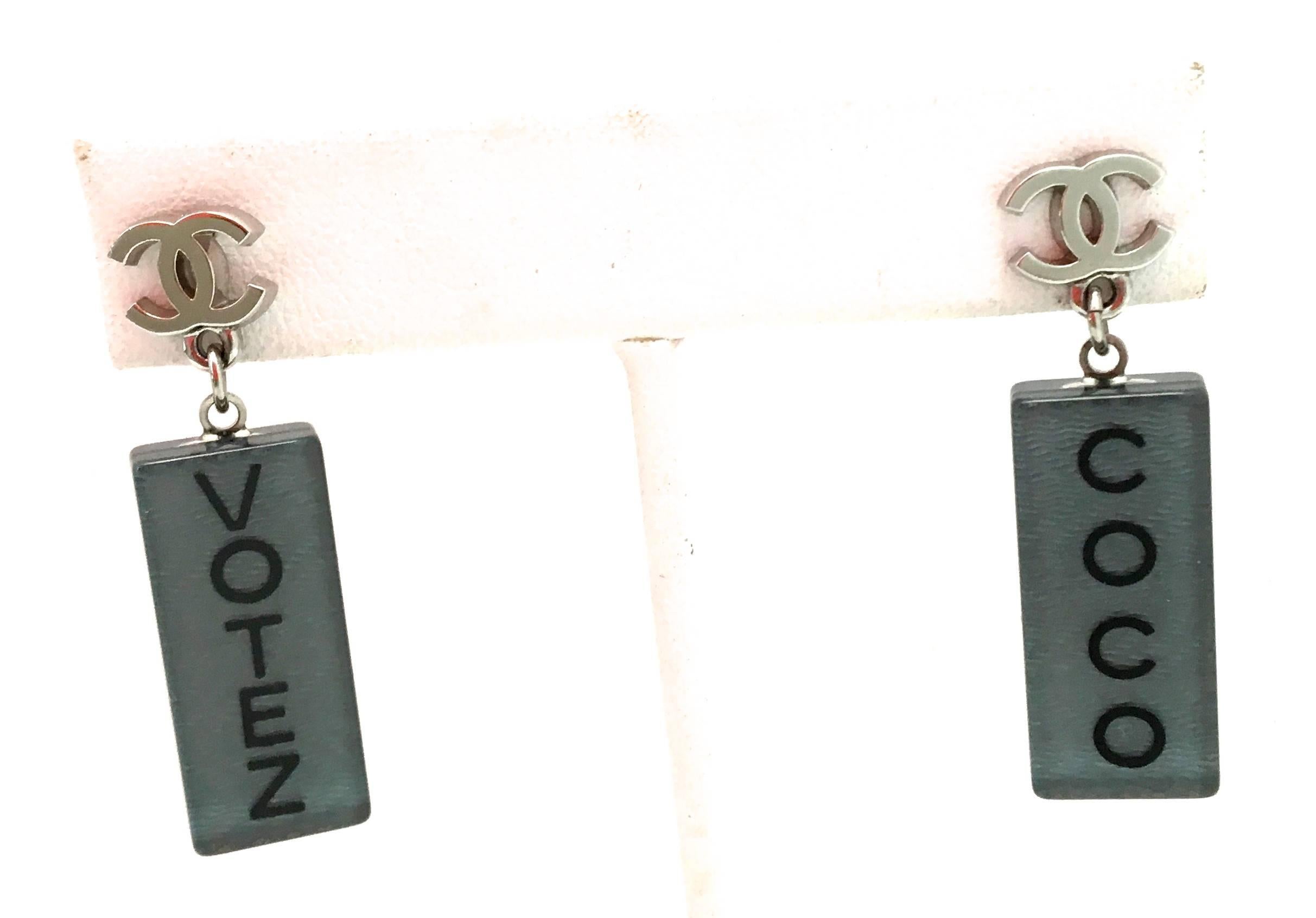 Presented here is a new pair of Chanel lucite earrings. Each earring has a silver tone CC logo with dangling lucite rectangles with a gray / blue color tone on either side of the earring. One earring says 'Votez' and the other says 'Coco.' The