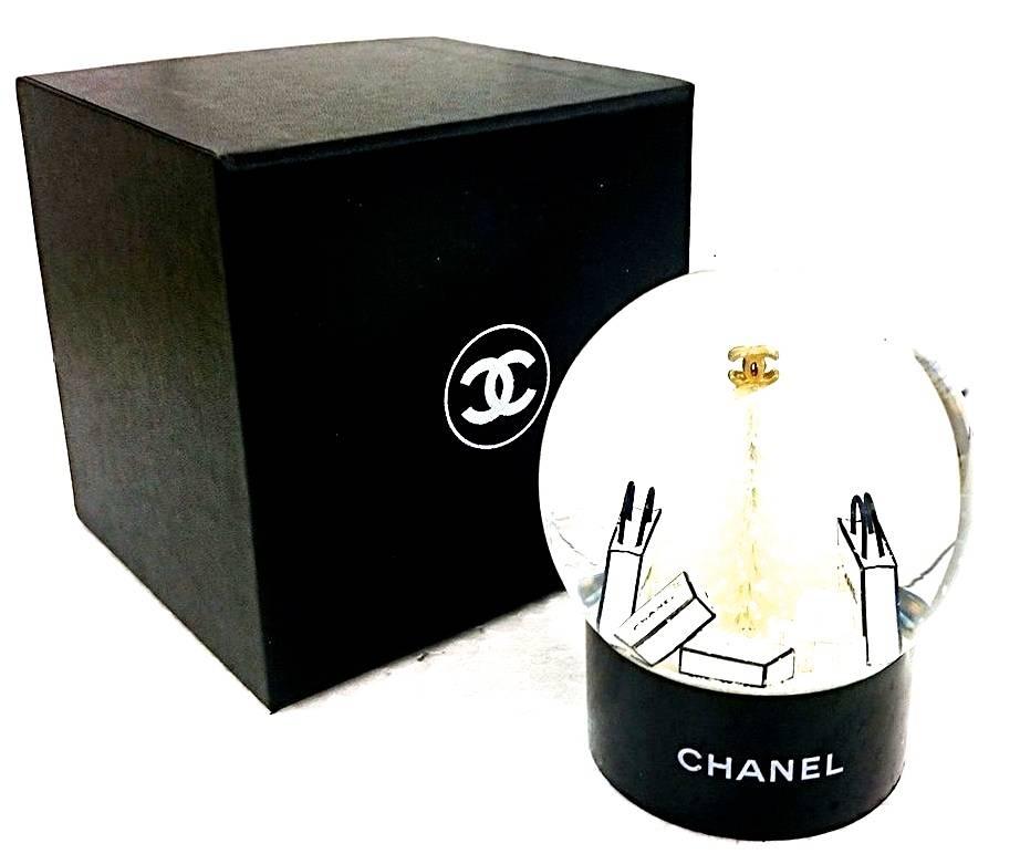 Presented here is a rare Chanel mini snow globe. The globe is comprised of a glass snow globe set atop a black base that is signed 'Chanel.' The snow globe has a white tree inside the glass surrounded by packages and bags that are signed 'Chanel.' 