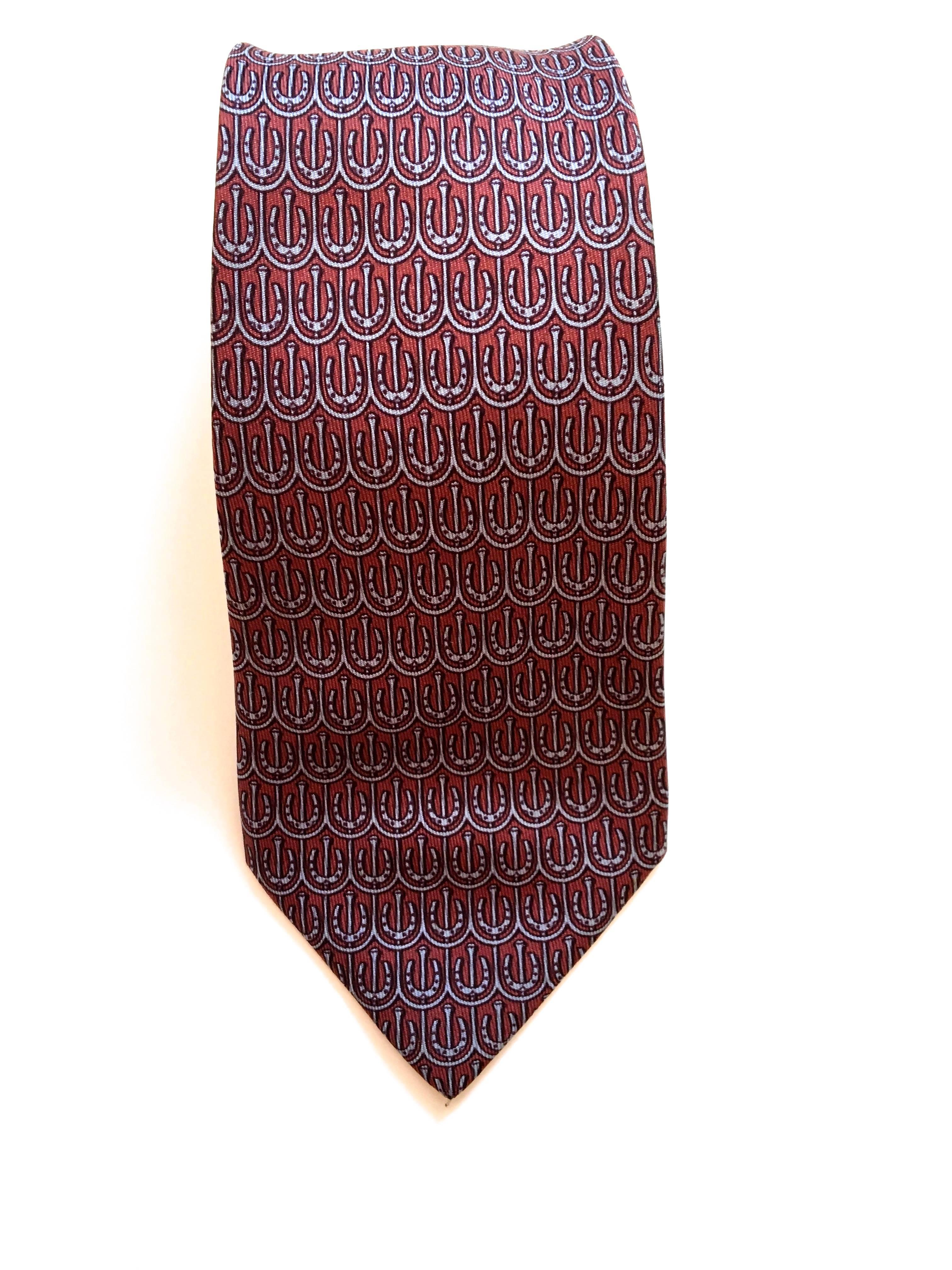 Presented here is a vintage silk necktie from Hermes. This beautiful tie is comprised of a background color of rust. In the foreground, there is a tessellation of indigo and gray horseshoes. The tie is 56 inches in length and 3.2 inches at the