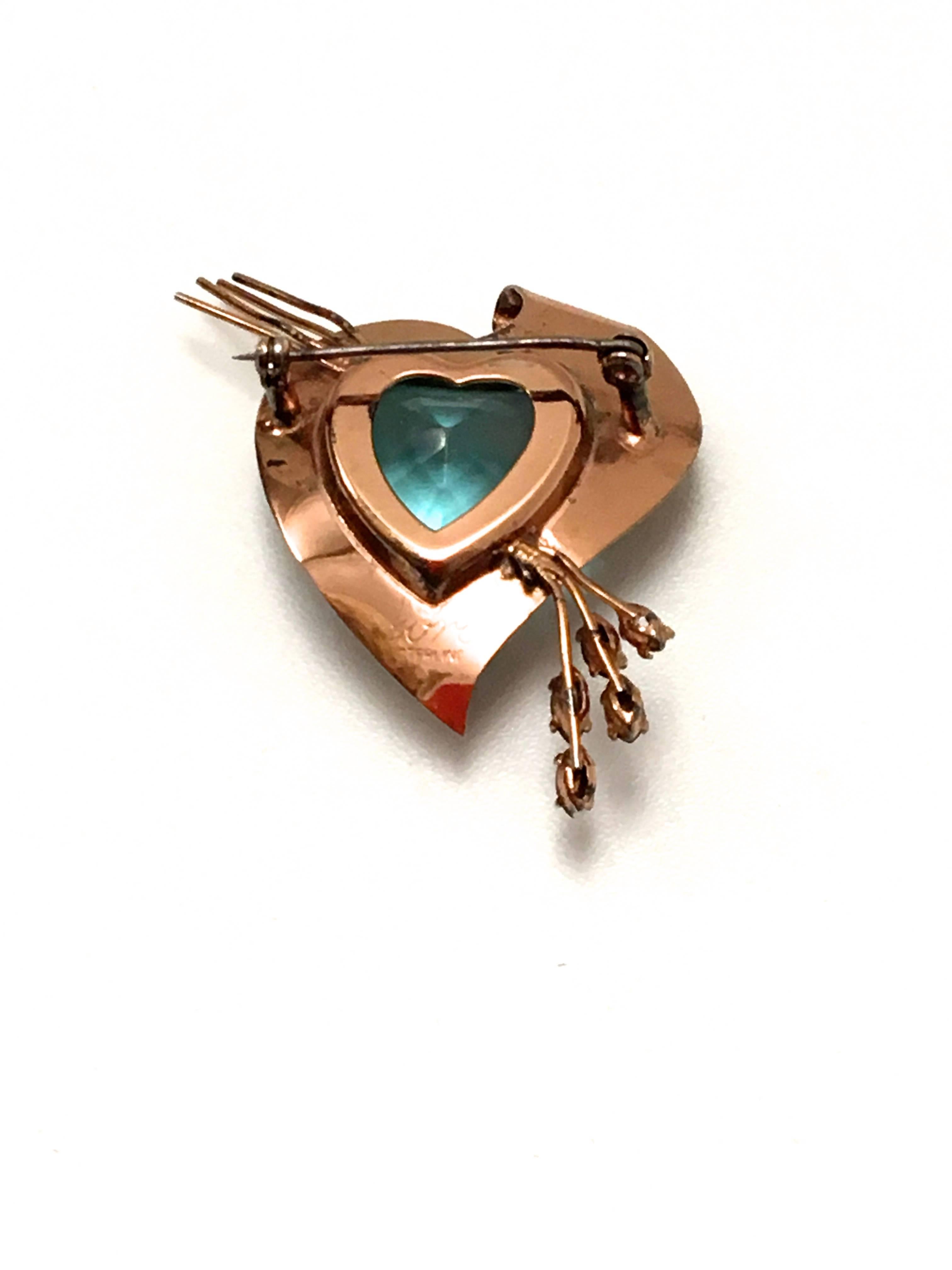 Presented here is a brooch from Coro from the 1950s. This beautiful brooch is comprised of a sterling silver covered with gold vermeil brooch frame. The main frame of the brooch houses a heart shaped center stone. The stone is a vibrant bright blue.