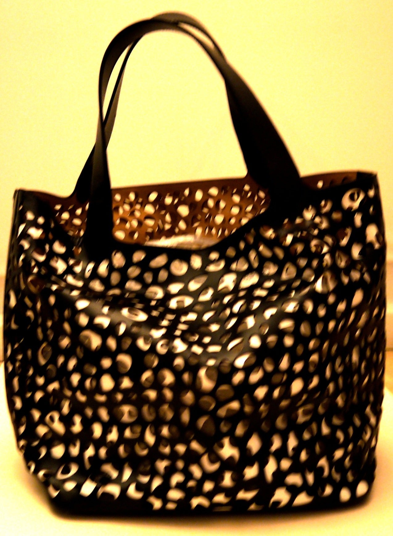 Gorgeous Alaia tote bag. A remarkable bag that has a layer of white leather with a holed pattern on the interior, and the exterior leather layer is a black leather with holes. The interior color of the bag is a beige. A gorgeous tote bag that makes