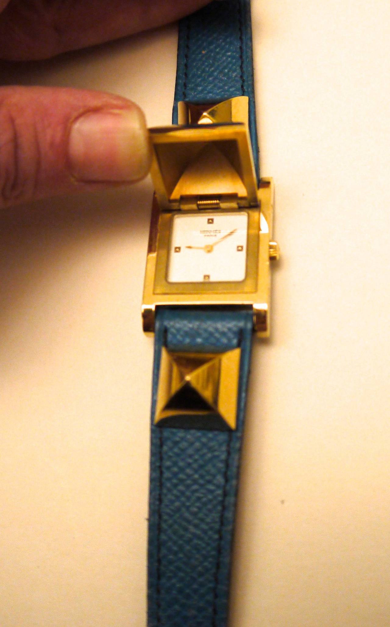 Vintage medor Hermes watch from the 1990's. The watch has a blue strap with gold tone hardware. The center gold square medor stud lifts up to reveal the watch face. The watch is a fantastic complement to any personal watch or Hermes collection.