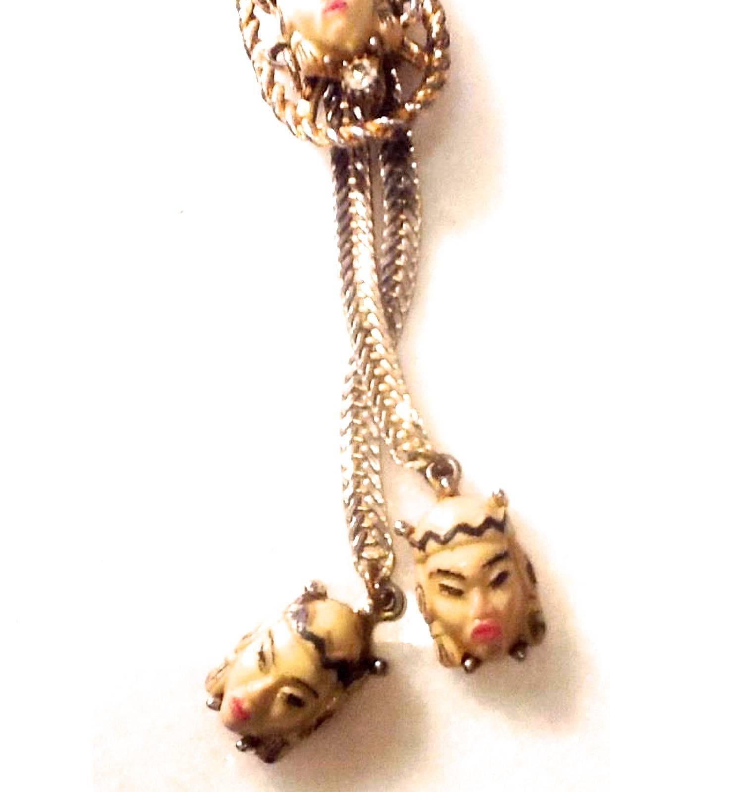 Presented here is a beautiful example of a Selro lariat necklace. The necklace has a tie of a female figure at the slide of the necklace. There is a charm on the end of each gold tone metal cord also with smaller Asian style heads. The slide of the