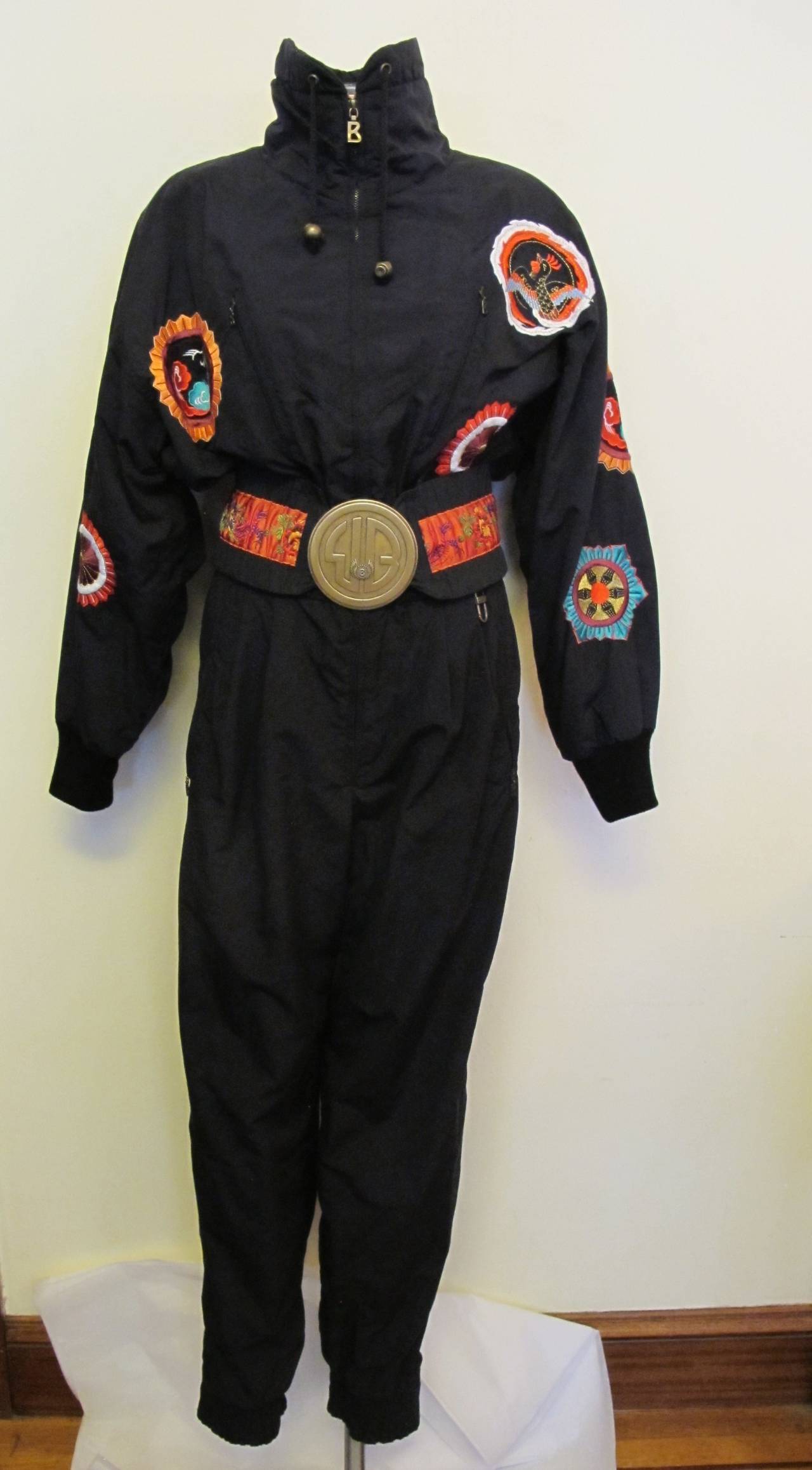 This exciting ski suit, gloves and purse were donated to Helpers by a San Francisco Grande Dame. There are 3 embroidered designs on the front of the jacket - 2 on each sleeve - and 3 on the back. The circular brass belt has the Bogner Logo which