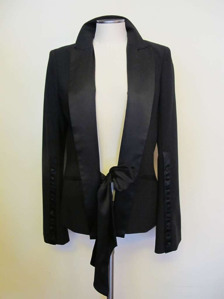 Chic black wool evening jacket with black satin labels and closure tie below the waist angled between two pockets. Pockets are bordered with satin. The sleeves are slightly bell shaped embellished by nine black satin buttons on each sleeve. Each