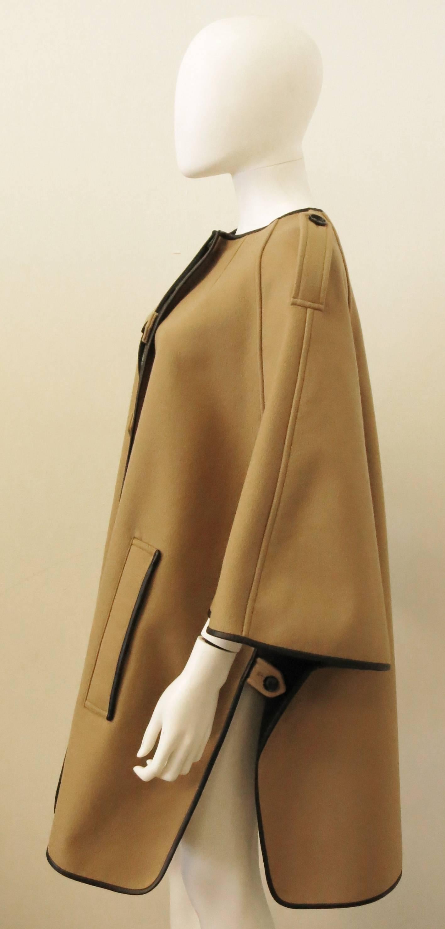A Burberry camel coloured cape coat made from a wool blend fabric with a dark brown leather trim.
The cape has soft rounded shoulders, seam detailing, epaulettes and a button fastening at the front. It also has two pockets and buttons at the side
