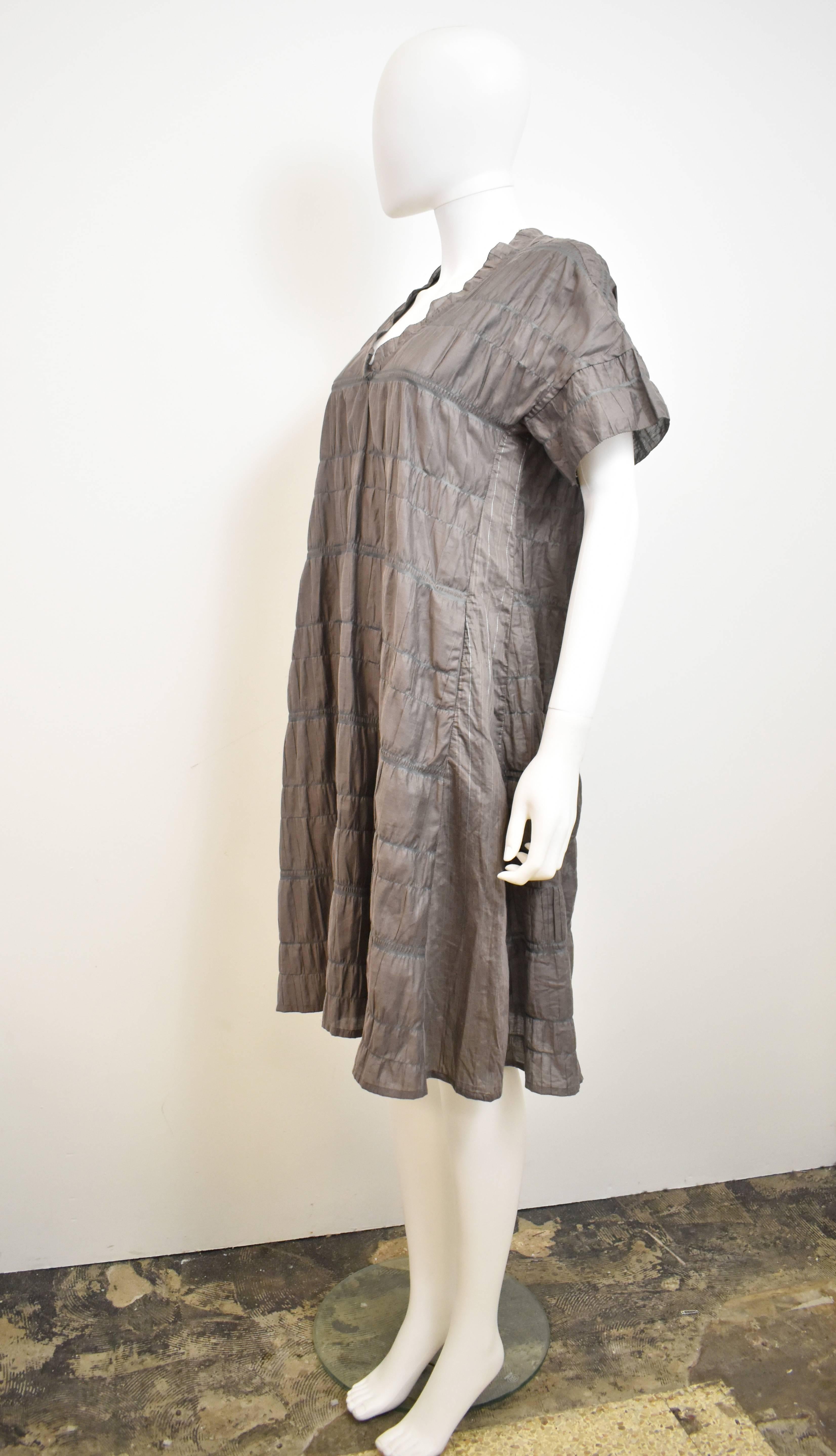 A grey pleated dress by Issey Miyake’s diffusion line Haat. The dress has a simple, loose shape, with V-neck, short sleeves and knee length skirt. The dress is made from a cotton and synthetic material blend which has been heat pleated vertically